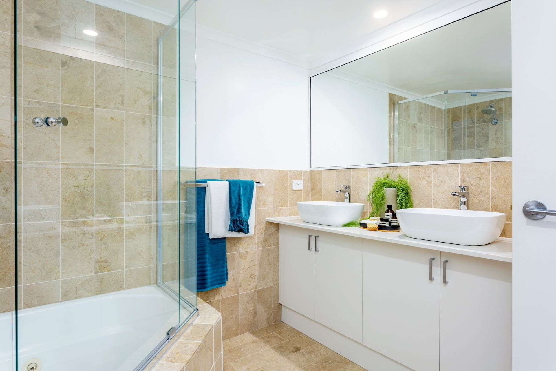 Bathroom with white vanity, vessel sinks, large mirror, and beige tile. Tub with glass shower door.
