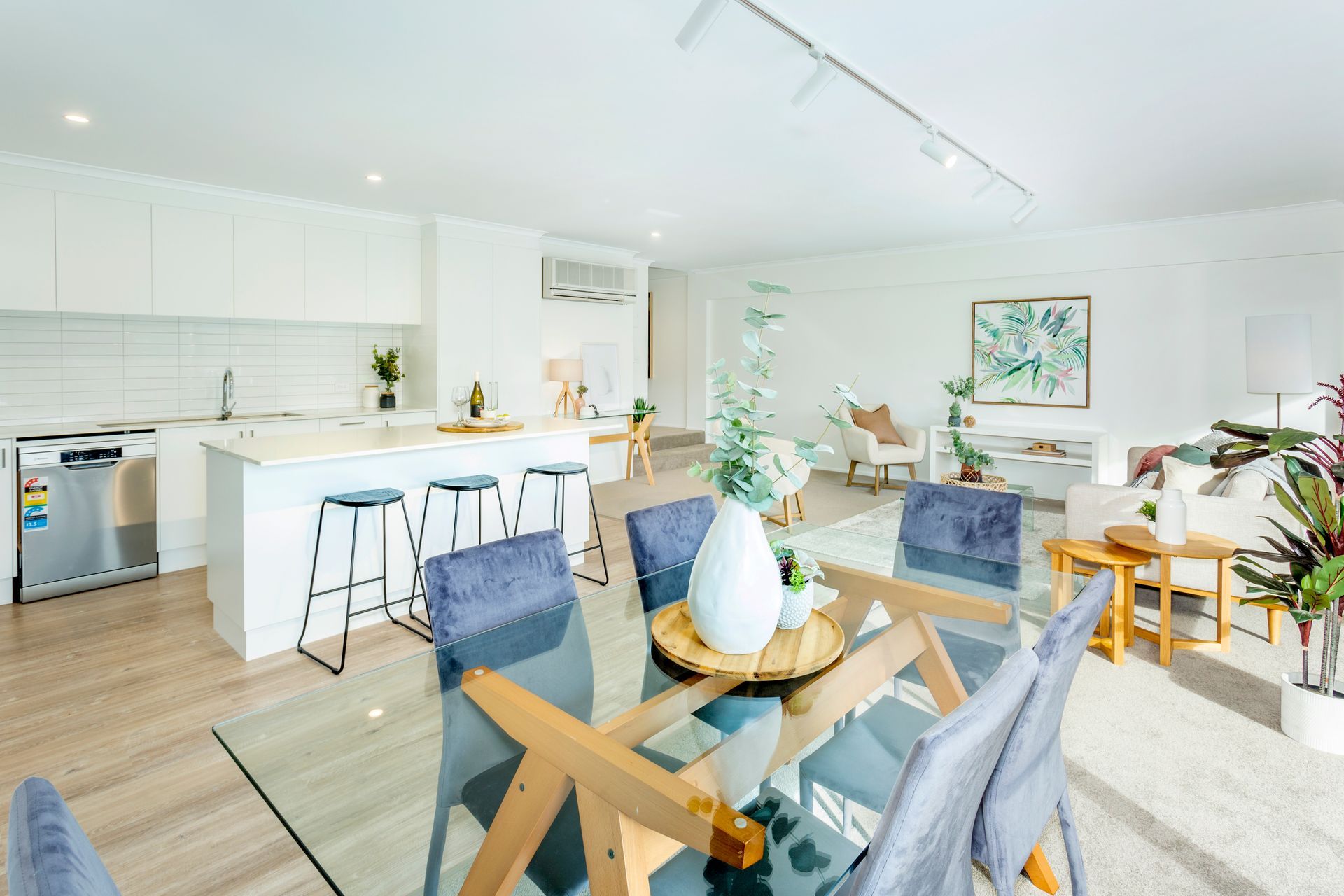 Open-plan living space with kitchen, dining area, and lounge. White walls, wood flooring, glass dining table, and blue chairs.