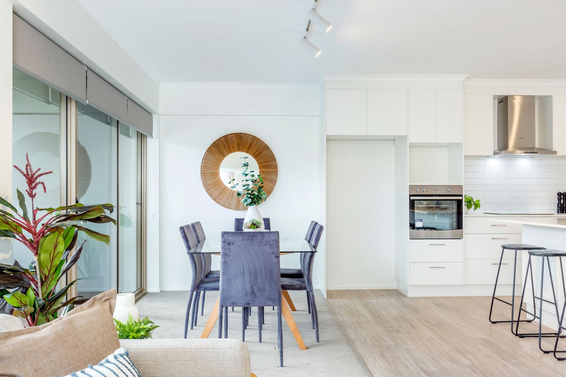 Dining area with table, chairs, mirror, and open kitchen with appliances; light wood floors, neutral tones.