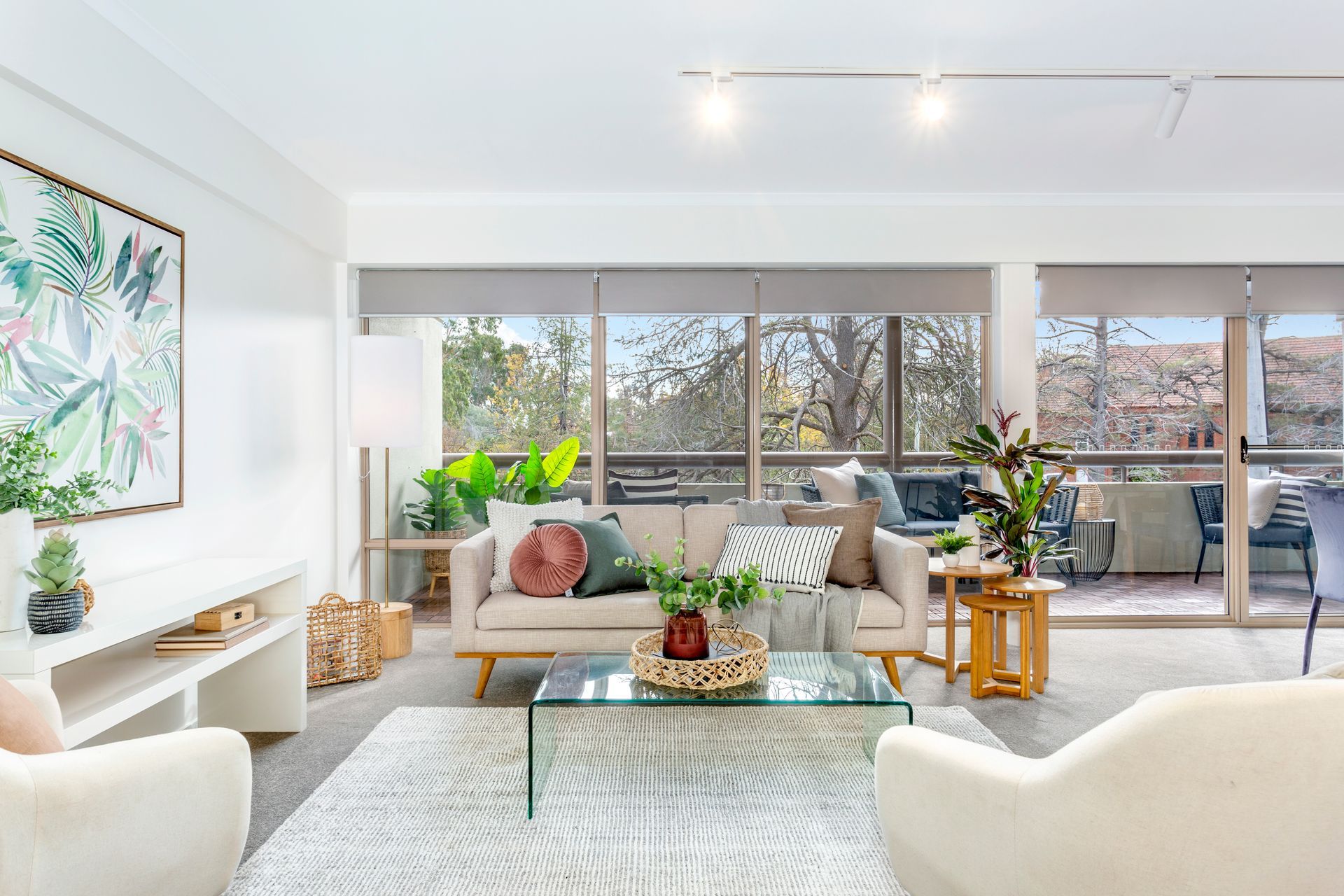 Living room with a beige sofa, two white armchairs, and a glass coffee table in front of a balcony.