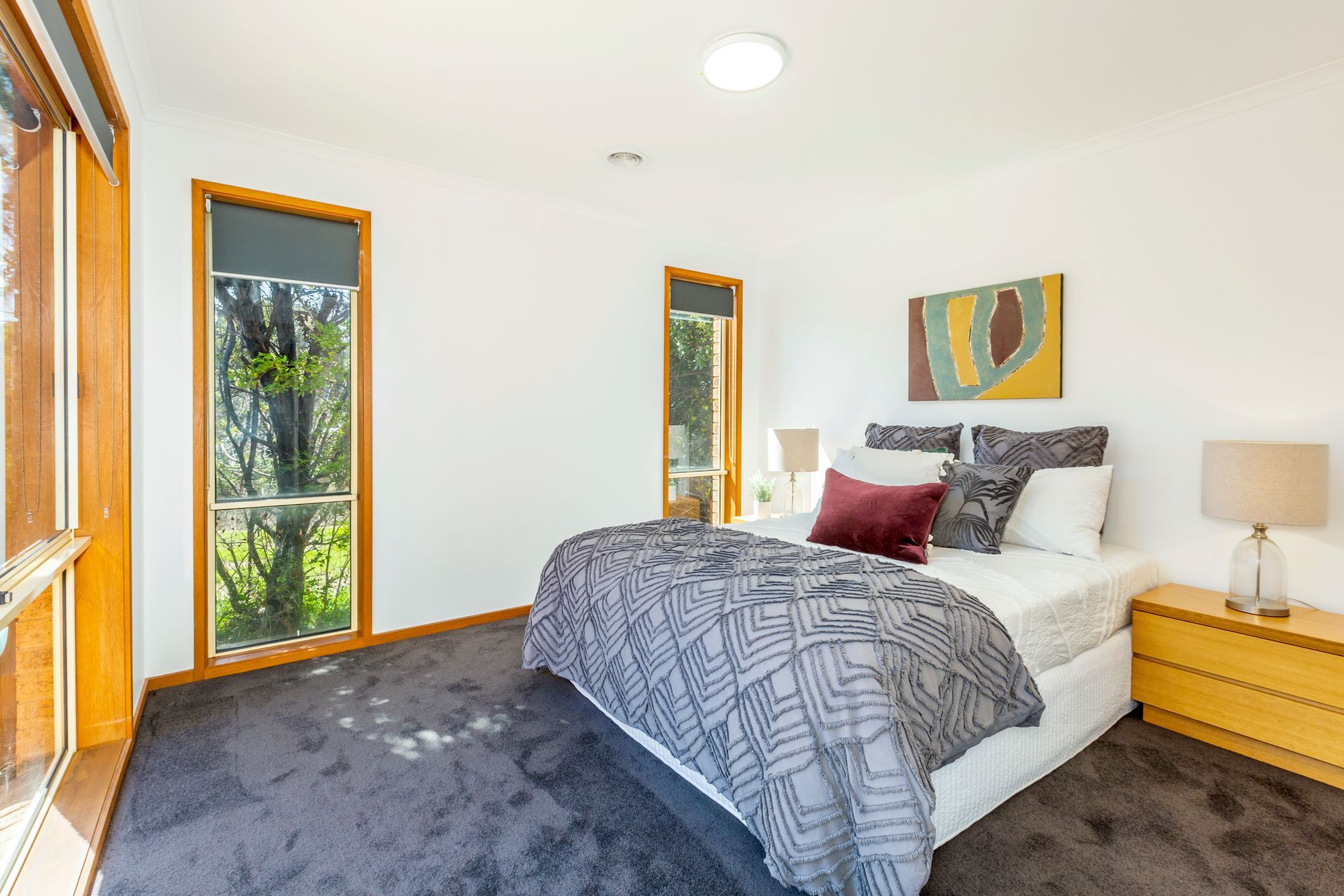 Bedroom with large windows, neutral walls, gray carpet, bed with decorative pillows and a colorful abstract art piece.
