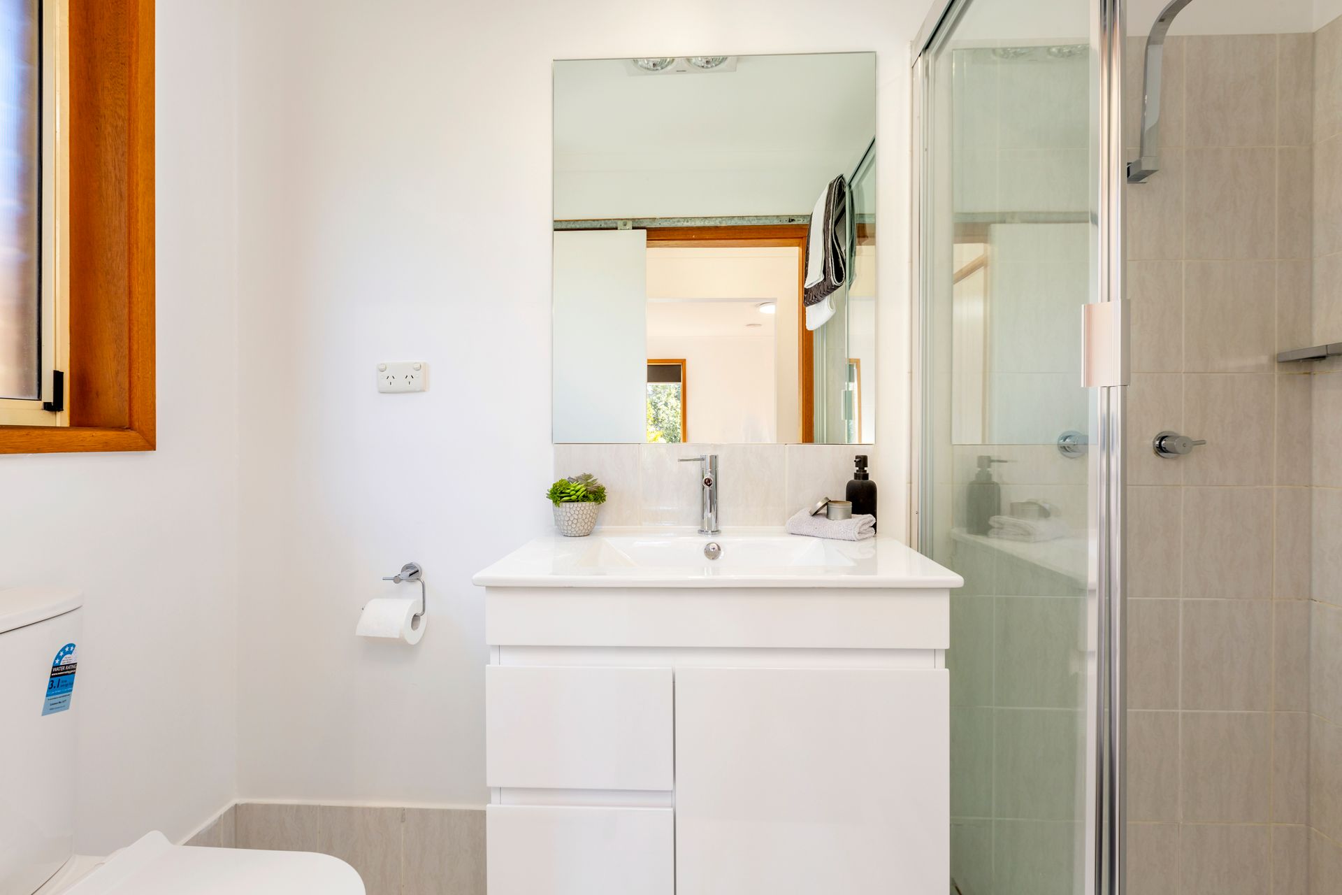 White bathroom with a vanity, mirror, and shower. Window on left.