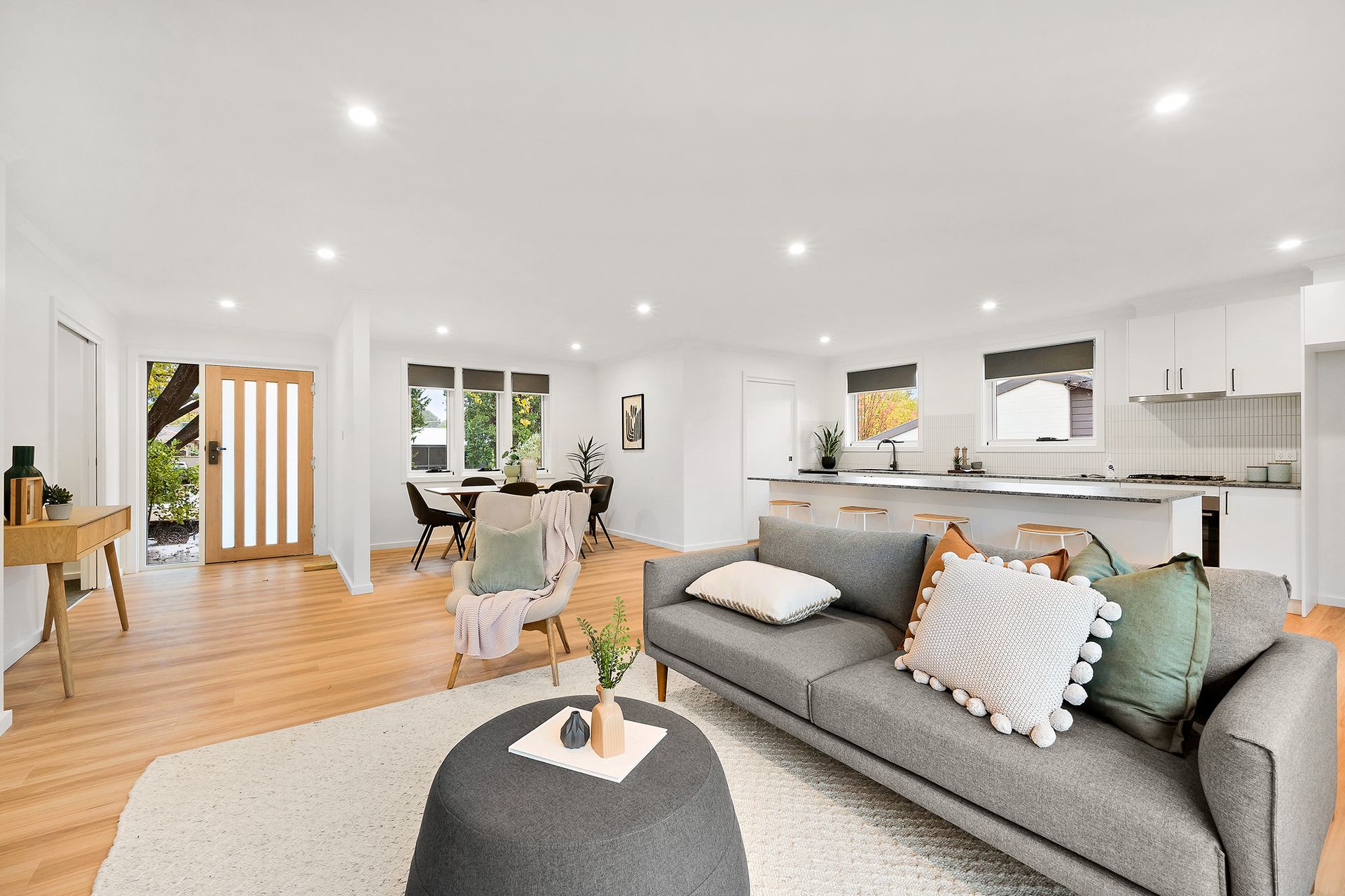 Open-plan living space with kitchen, dining, and living areas; features light wood floors, gray sofa, and white walls.