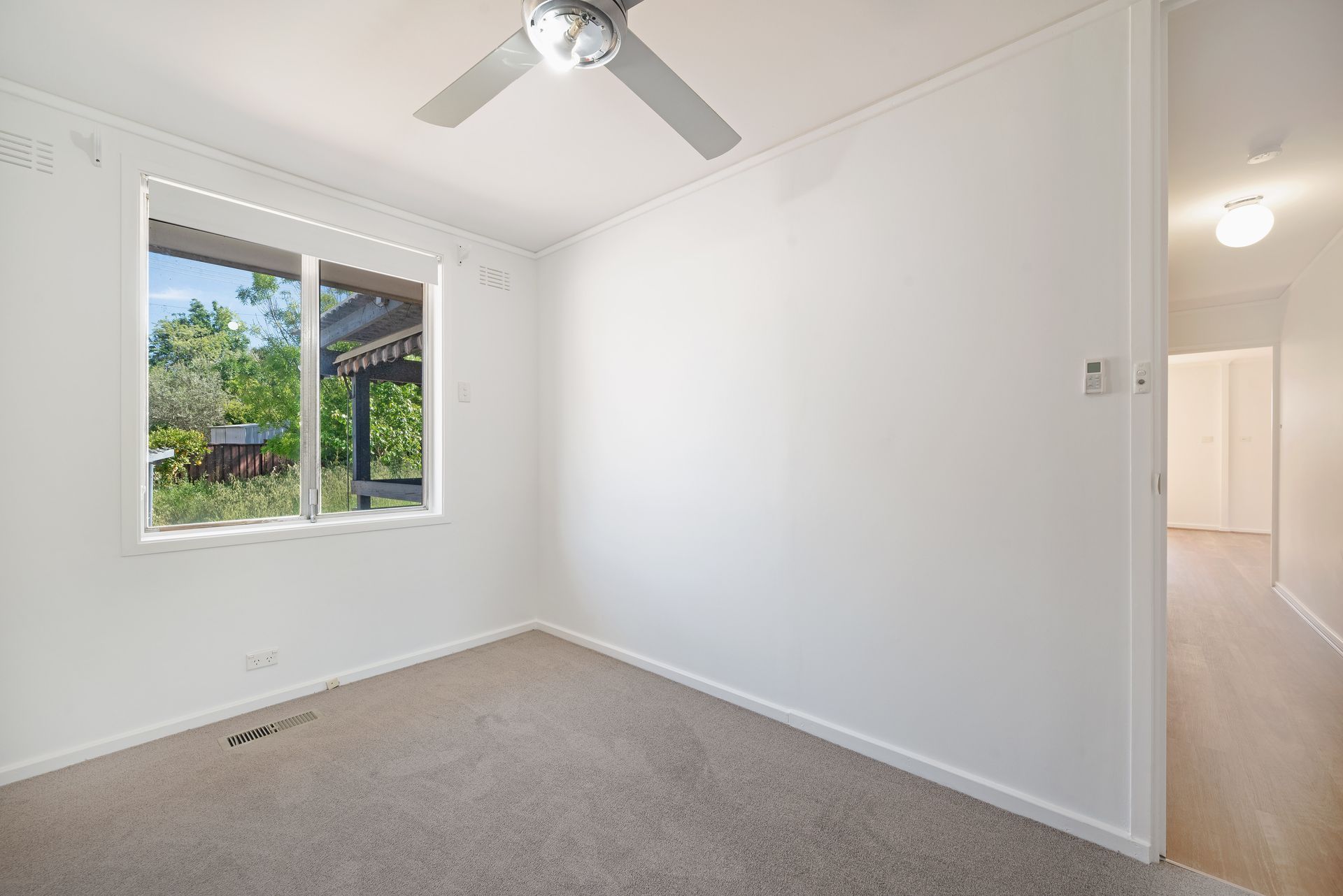 Empty room with gray carpet, white walls, and a window overlooking greenery. A hallway is visible to the right.