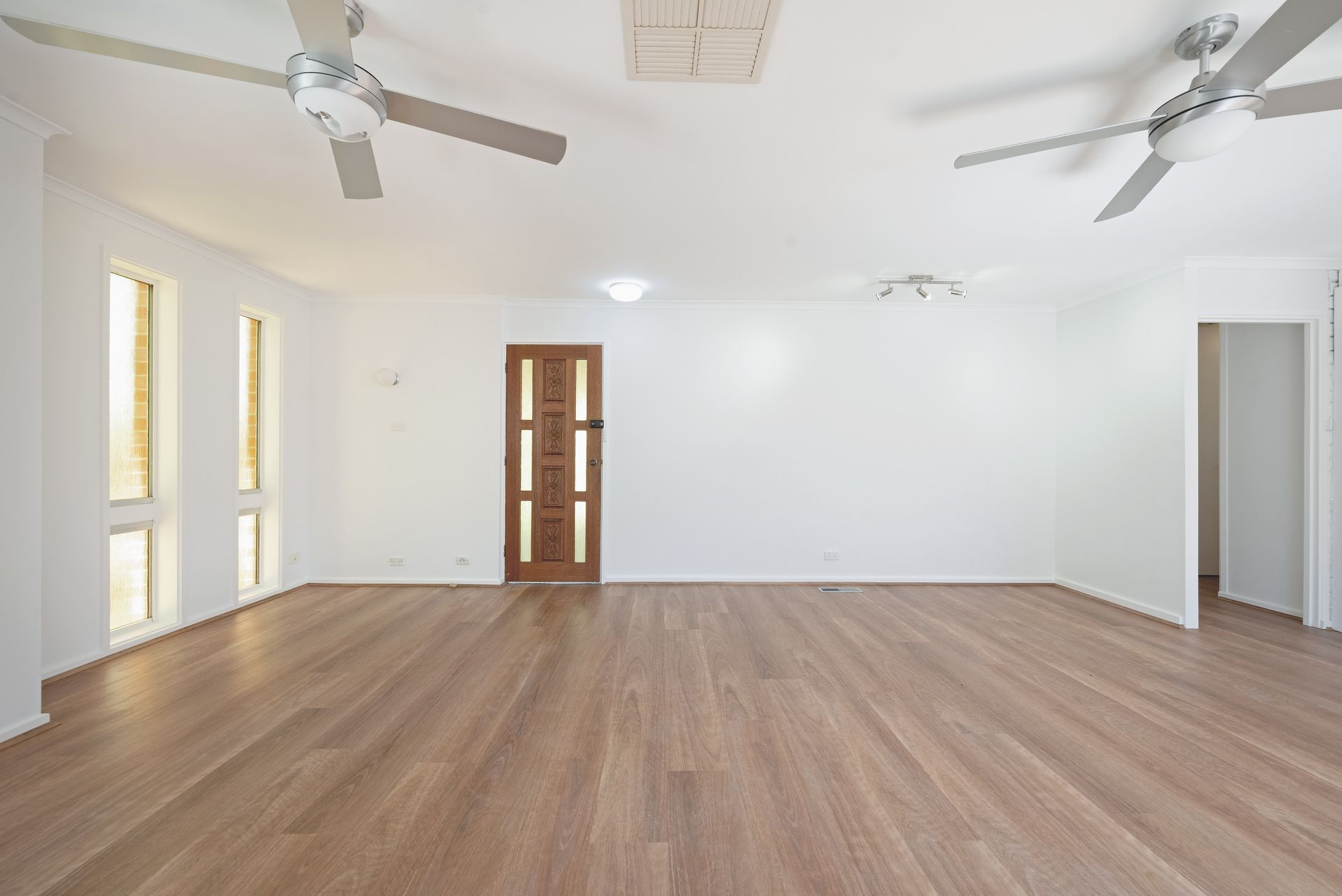 Empty room with wooden floor, white walls, two ceiling fans, and a brown door.