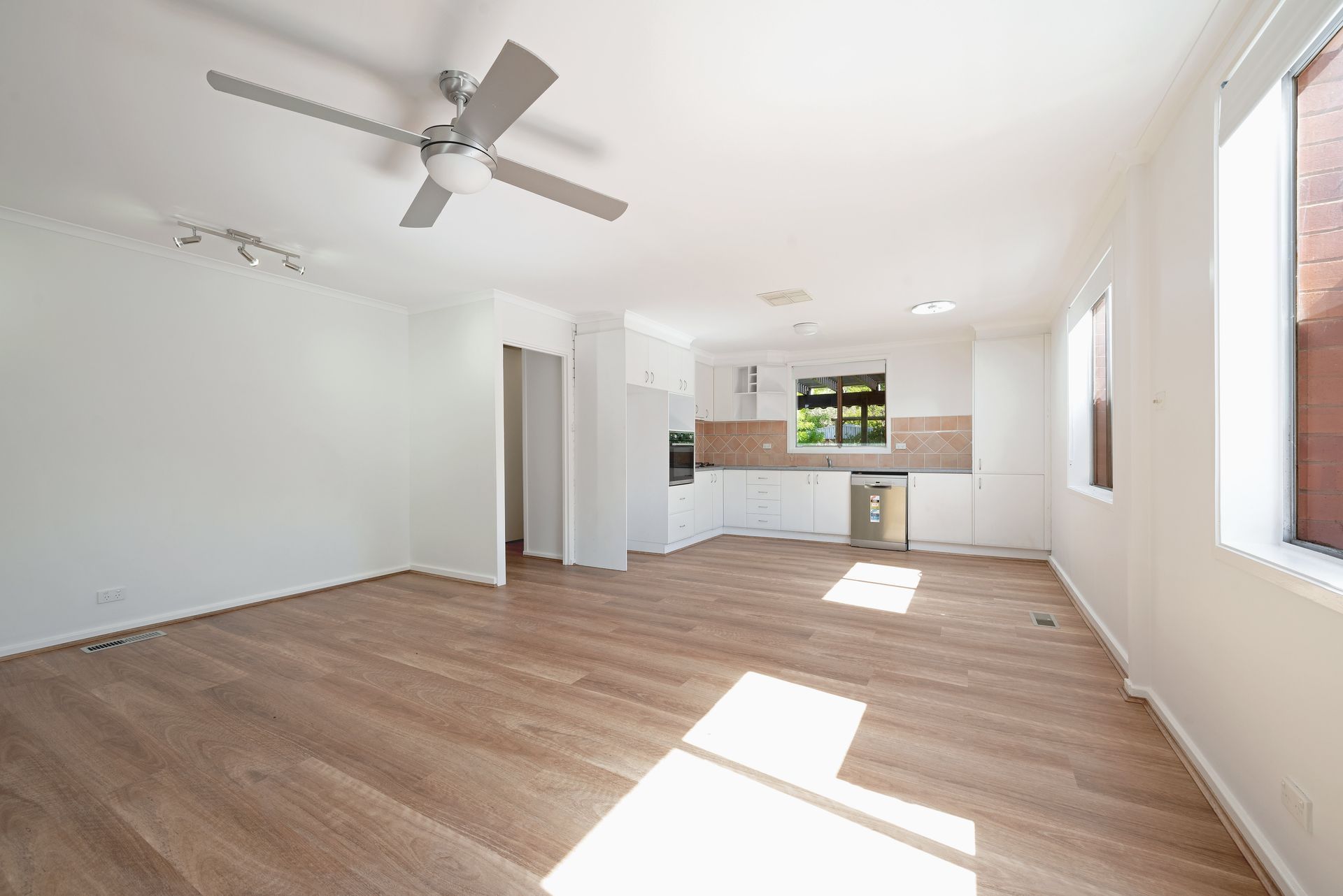 Empty, bright, open-plan room with wooden floors, white walls, kitchen, windows, and ceiling fan.