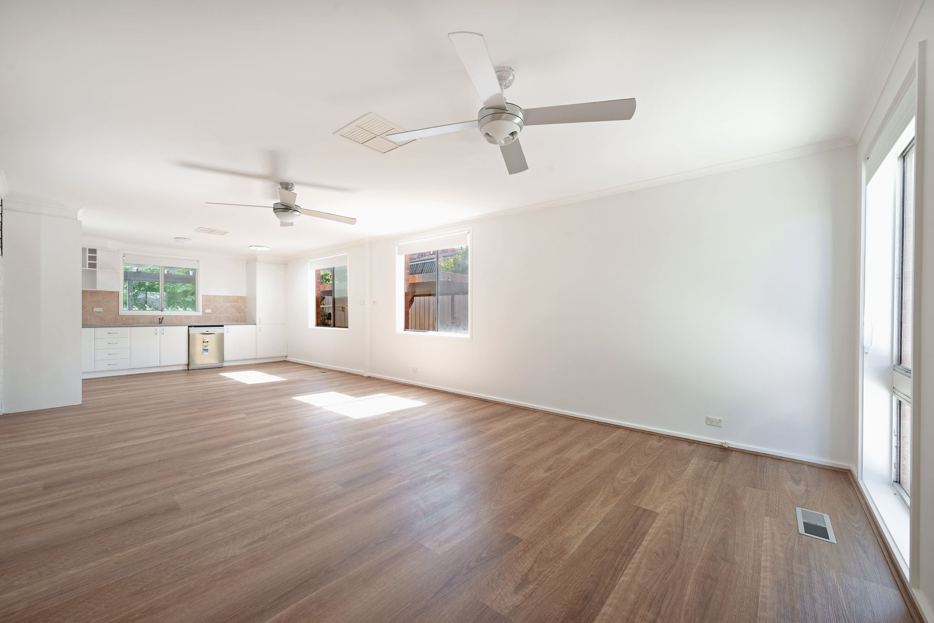 Empty room with wooden floor, white walls, two ceiling fans, and windows with sunlight.
