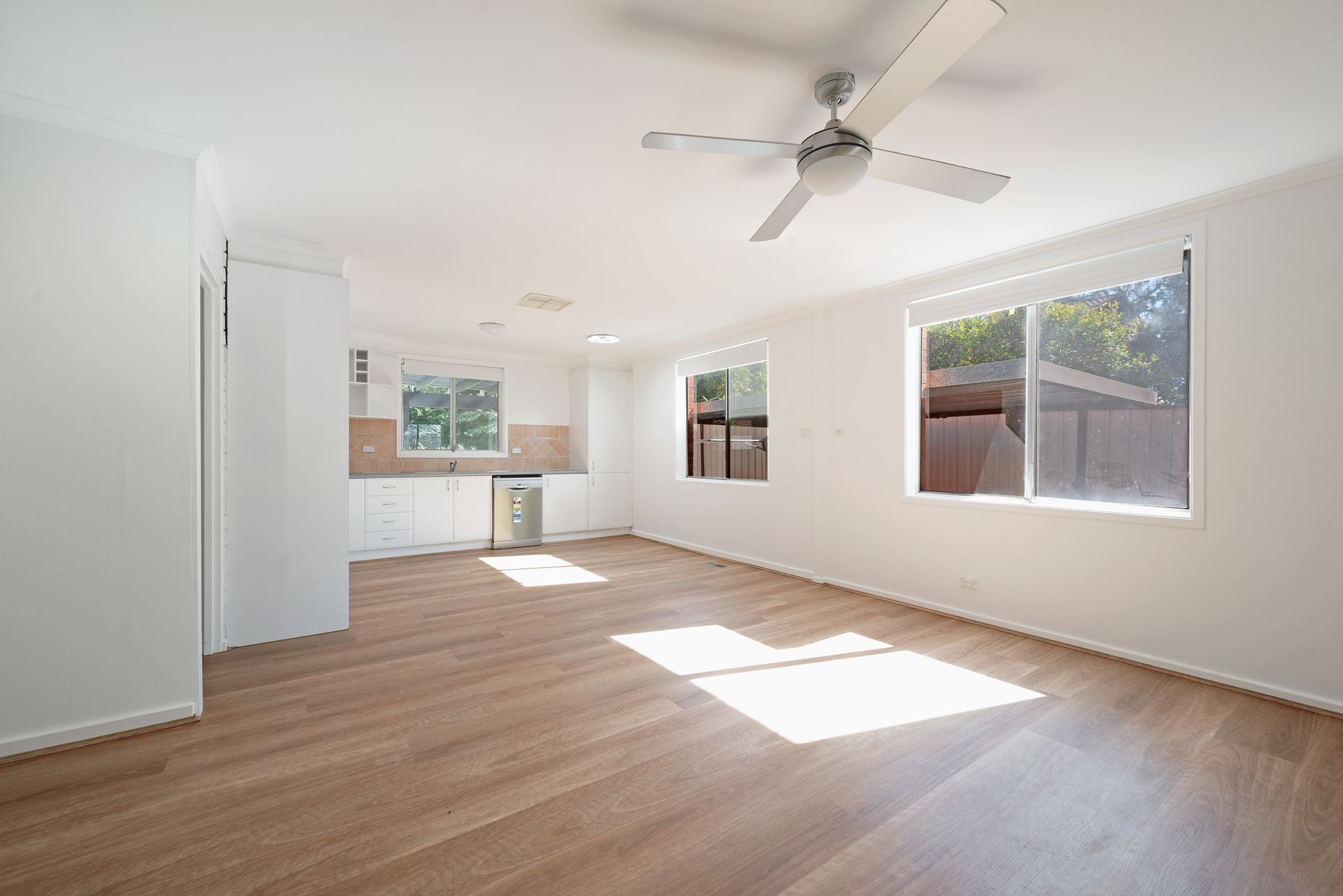 Bright, empty interior with wood floors, white walls, and a small kitchen visible. Sunlight streams through windows.