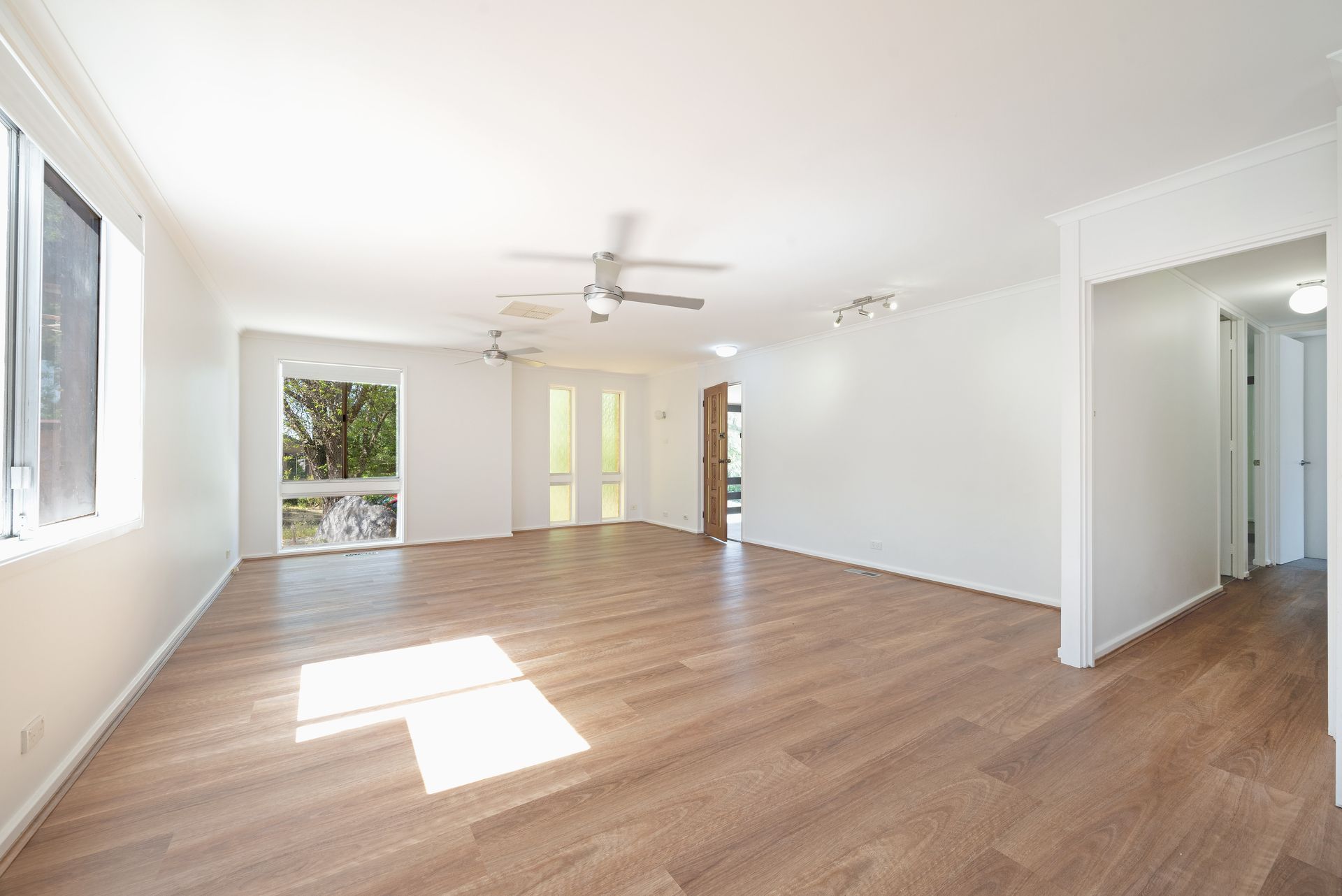 Empty, bright living room with wood flooring, white walls, and two ceiling fans.