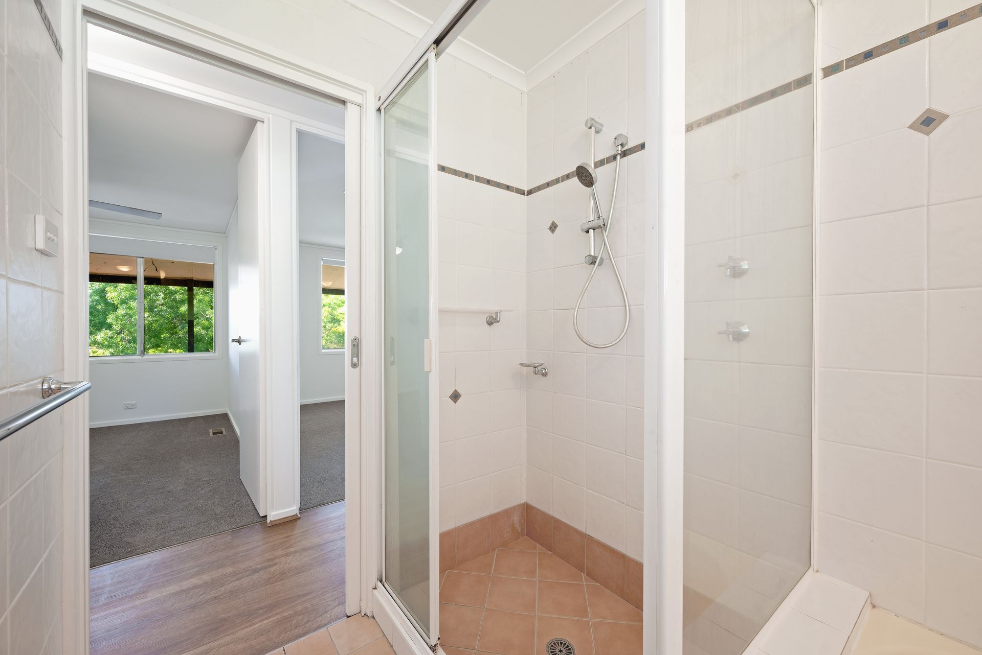 Bathroom with shower, doorway to a bedroom with window. Tile and glass accents.