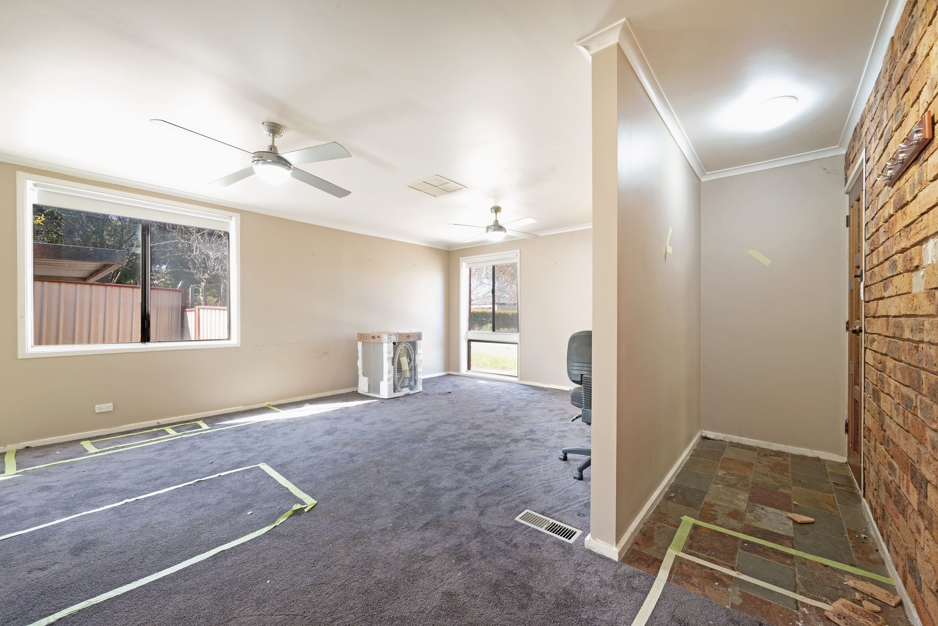 Empty room with grey carpet, beige walls, and brick accent. Ceiling fans and window visible.