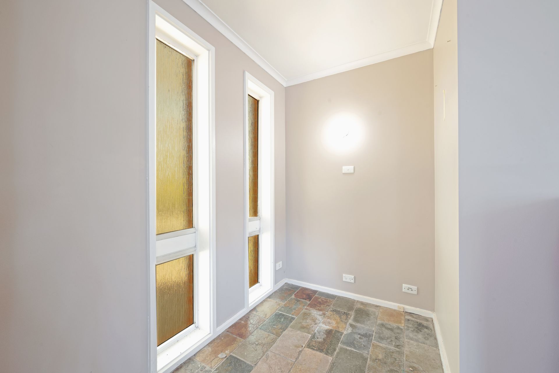 Narrow, empty hallway with two tall windows, textured glass, light gray walls, and stone floor.