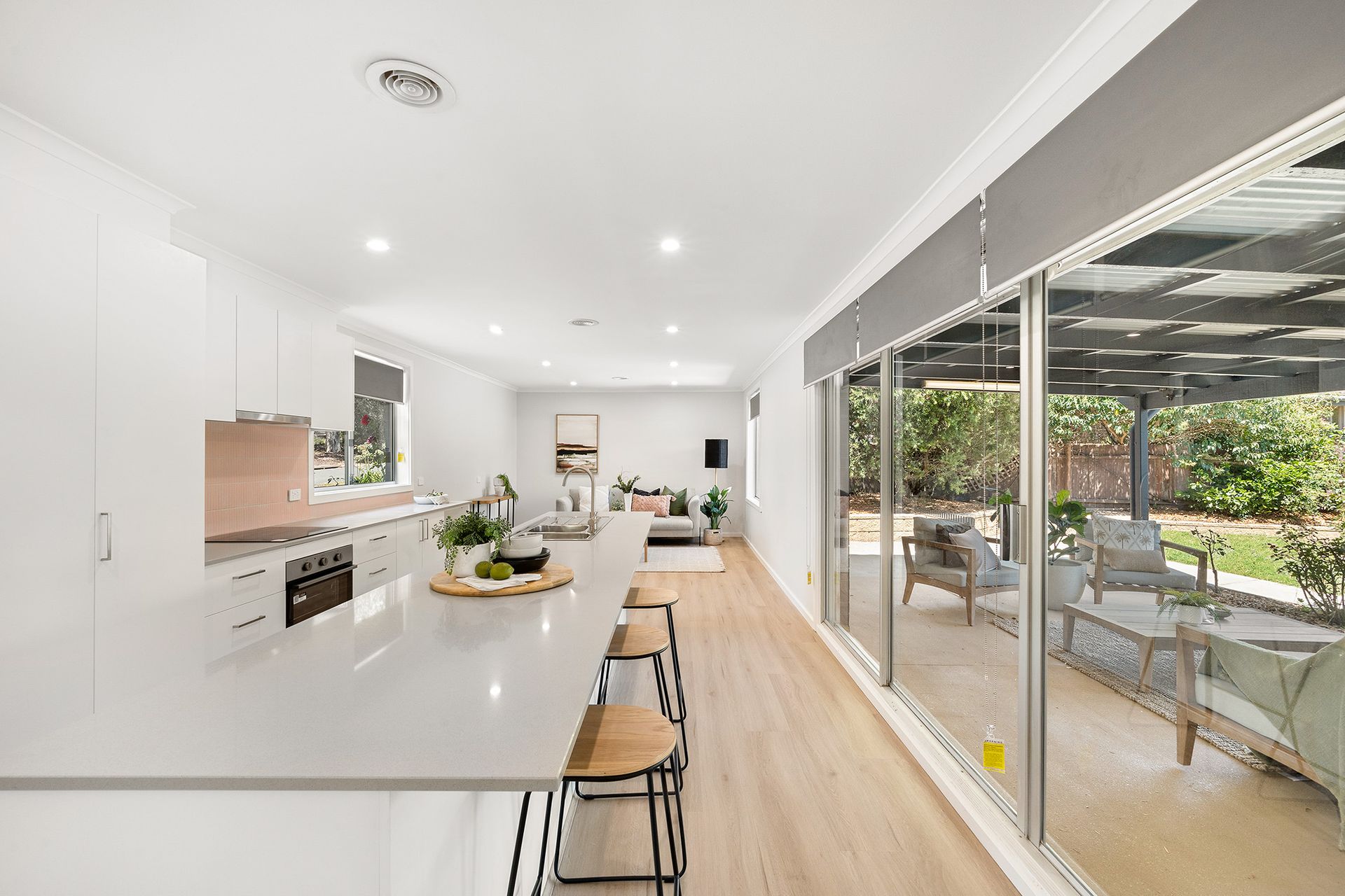 Modern kitchen and living area with long island, light wood floors, and large windows leading to an outdoor seating area.