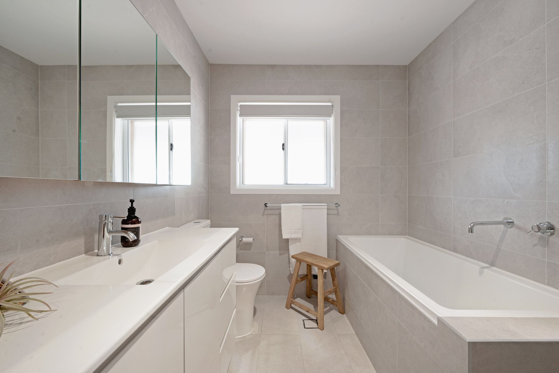A modern bathroom with a white sink, tub, and toilet. Light gray tiles cover the walls. A small wooden stool stands near the tub.