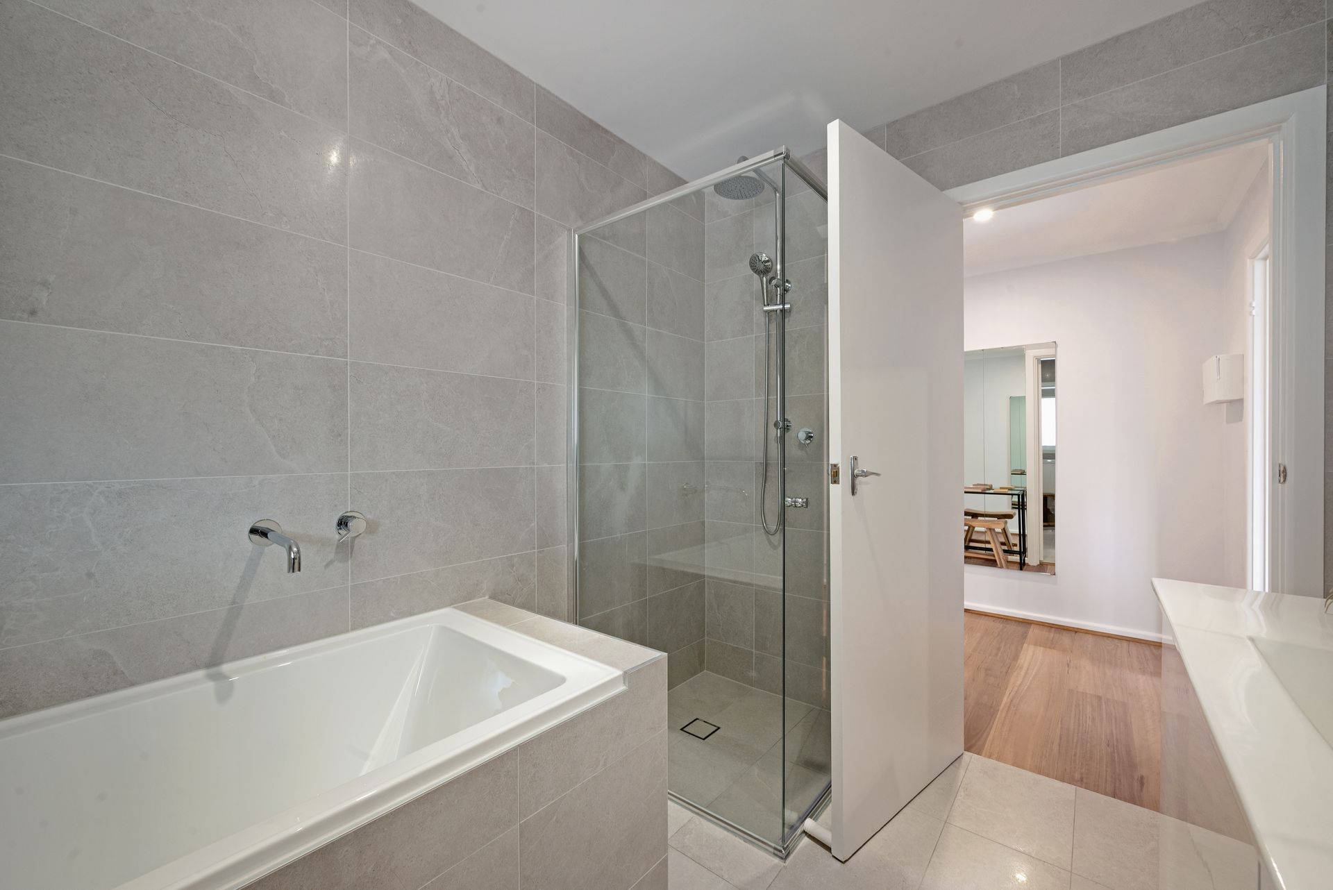 Modern bathroom with a bathtub, glass shower, and open doorway to a hallway. Gray tiled walls and floor.