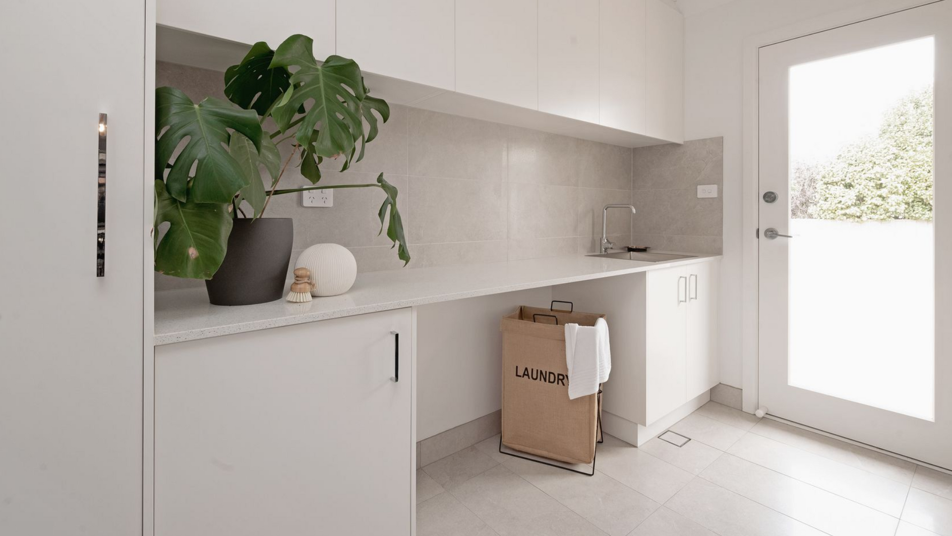 Laundry room with white cabinets, countertop, and door. A Monstera plant sits on the counter next to a laundry bag.