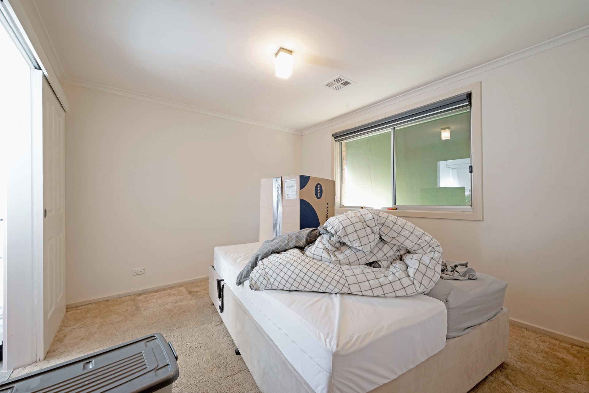 Empty bedroom with a bed, boxes, and a window. Beige walls and carpet.