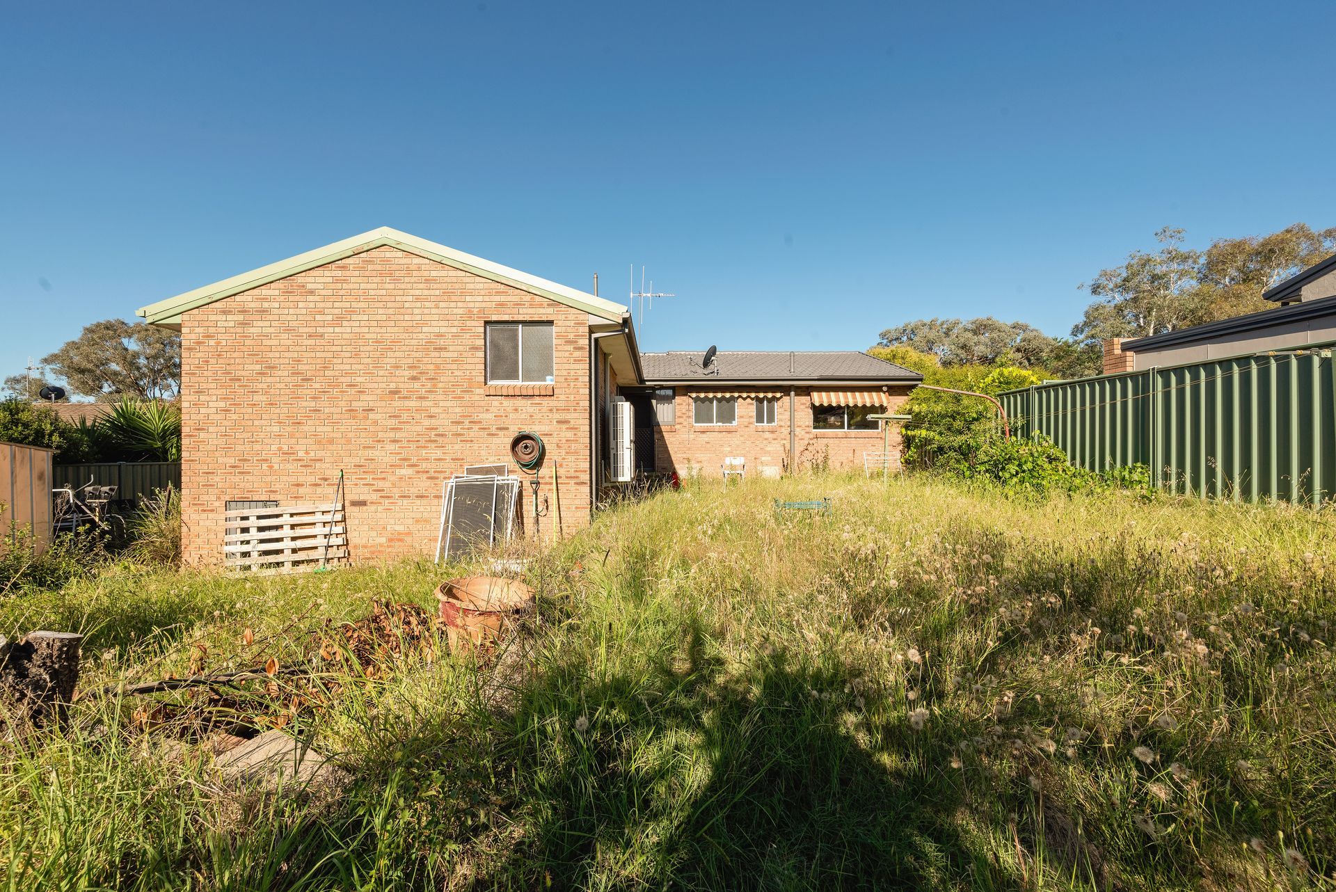 Overgrown backyard of a brick house under a blue sky, with tall grass and a green fence.