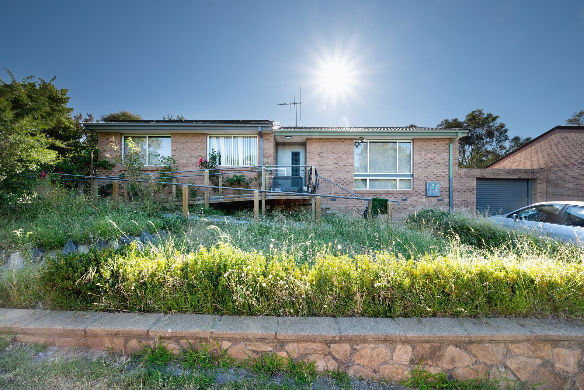 Brick house with overgrown grass in front, bright sun in the sky.