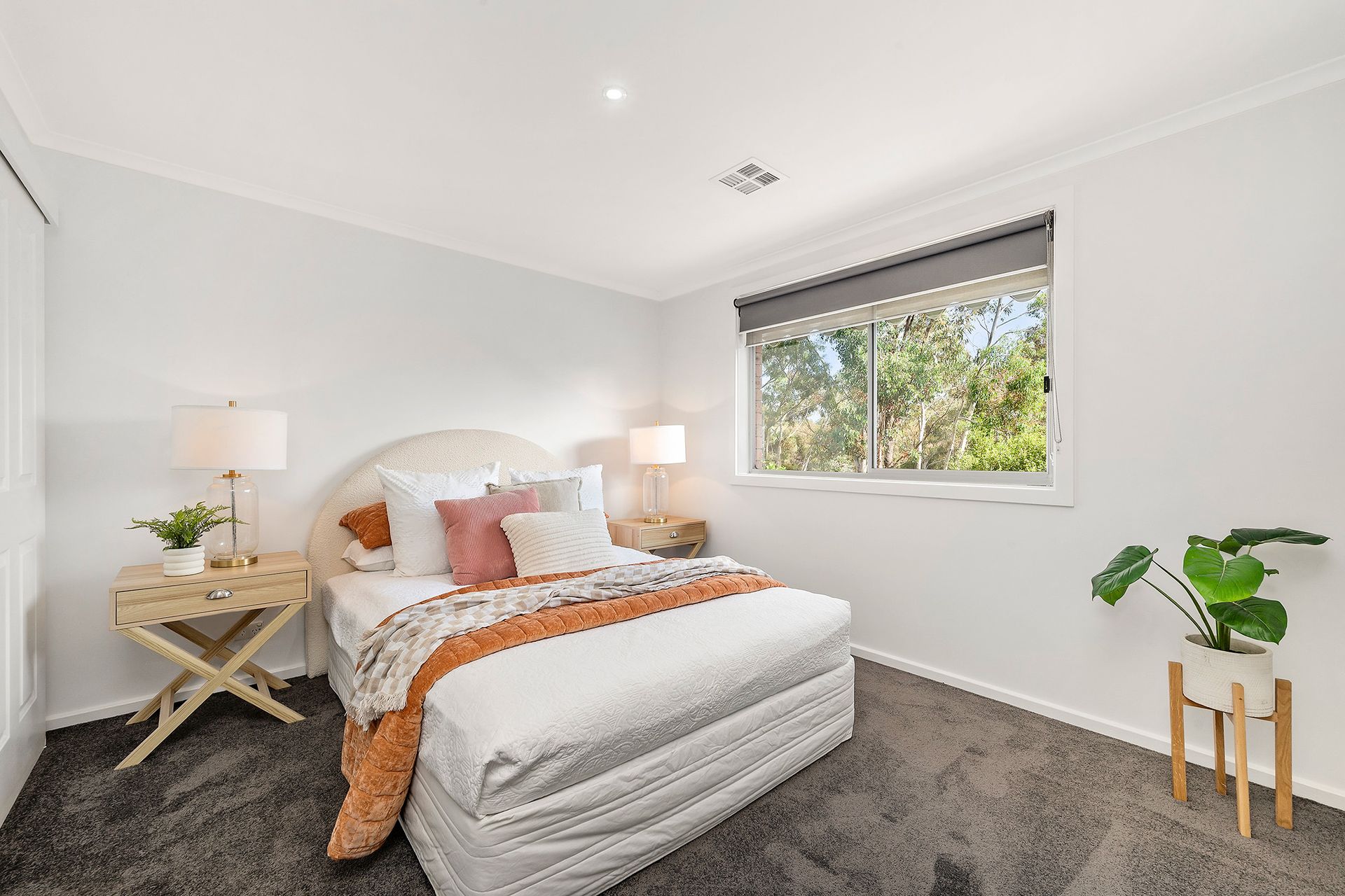 Bedroom with white walls, window, bed with orange and pink accents, two bedside tables, and a potted plant.