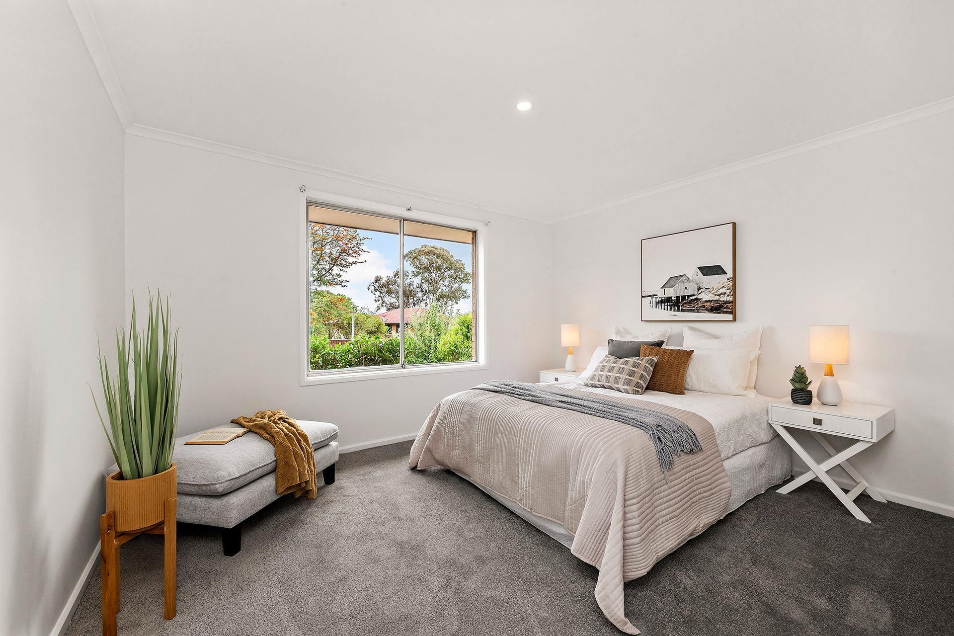 Bedroom with white walls, window, bed with orange and pink accents, two bedside tables, and a potted plant.