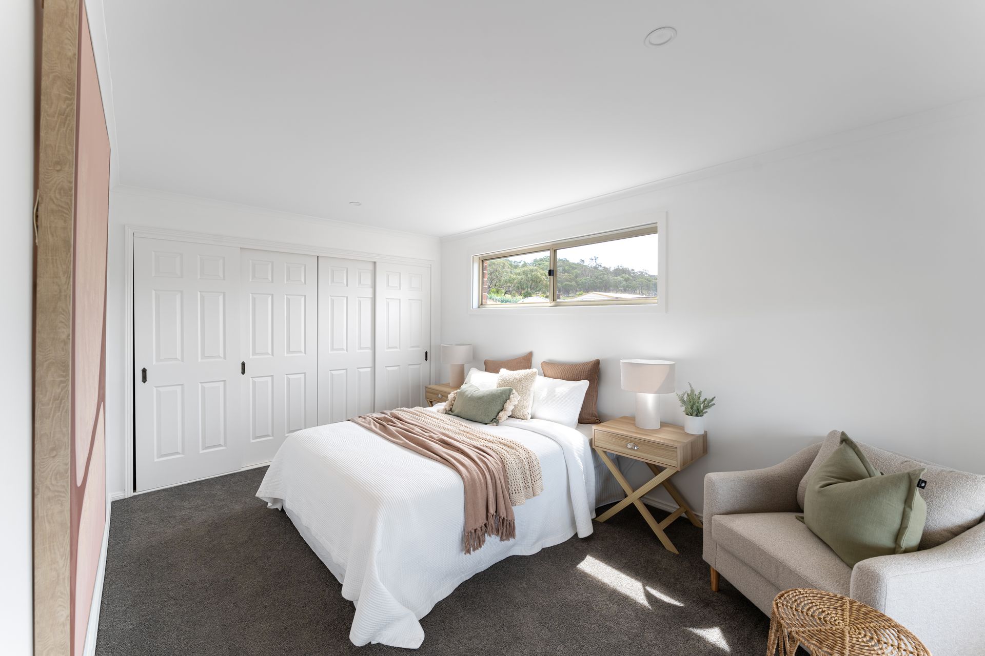 Bedroom with white walls, closet doors, and bed. Includes a neutral-toned chair, side table, and window.