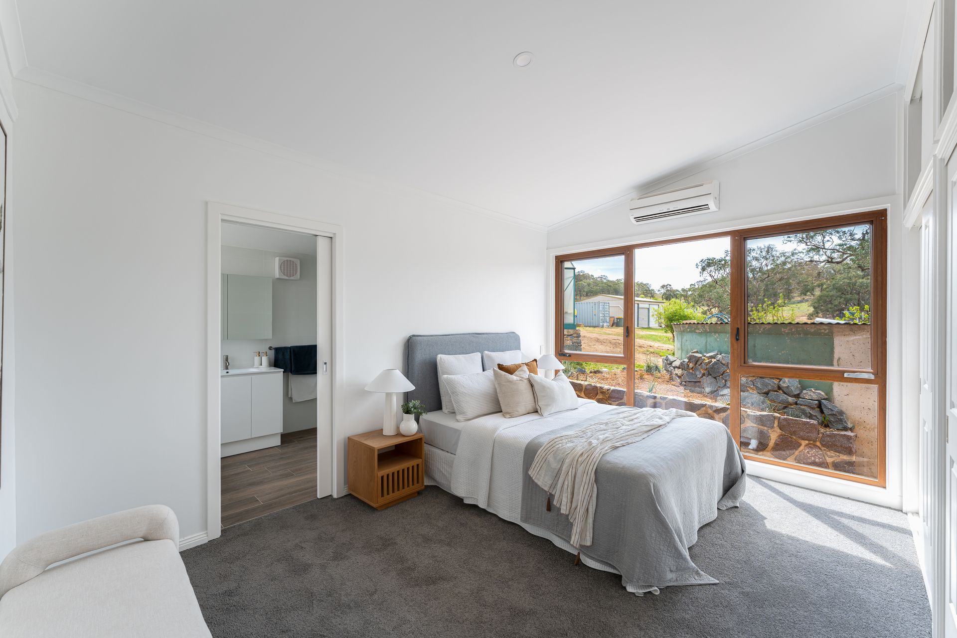 Bedroom with a bed, nightstand, and large windows looking outside; a door leads to a bathroom.