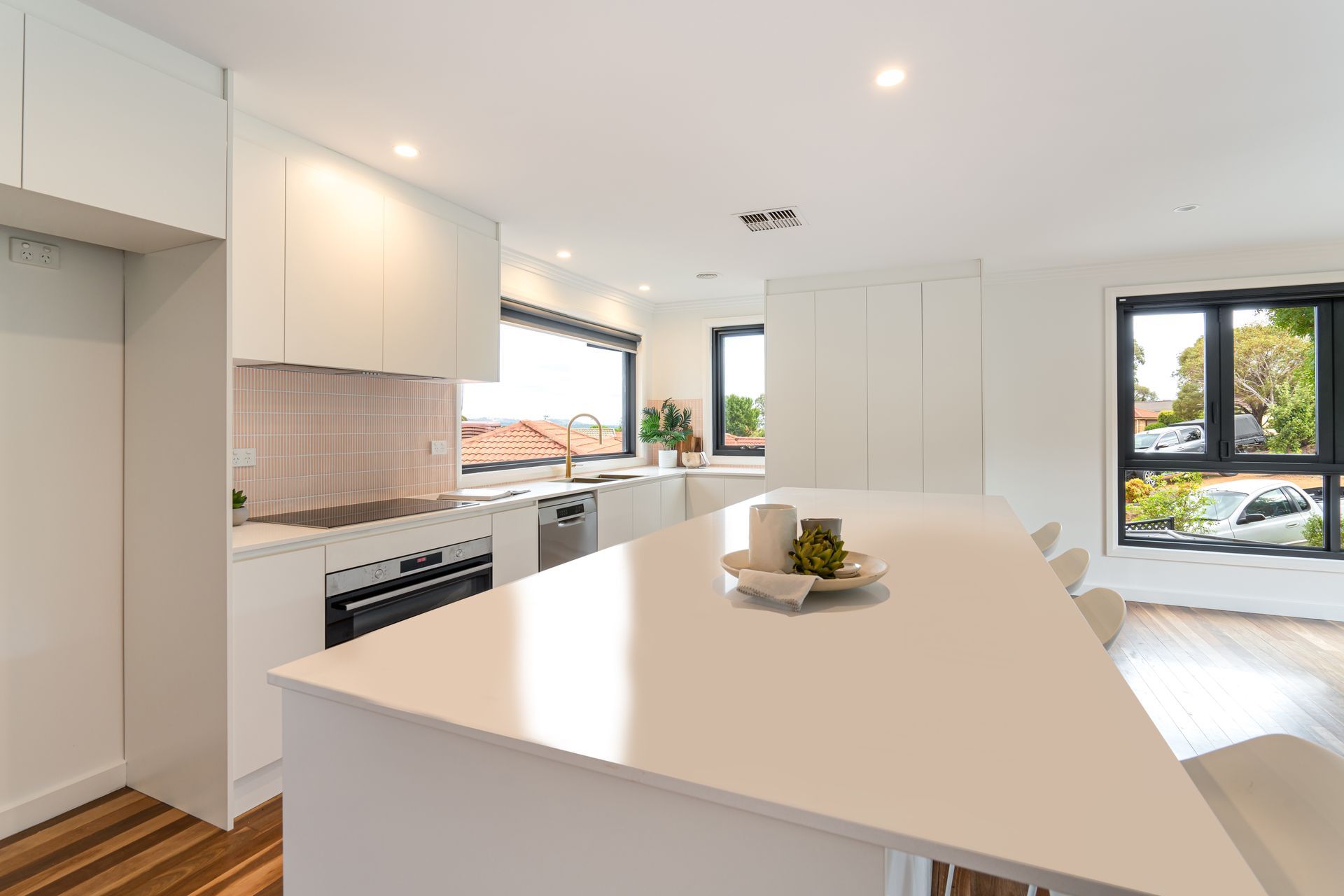Modern white kitchen with island, wooden floors, stainless appliances, and windows.
