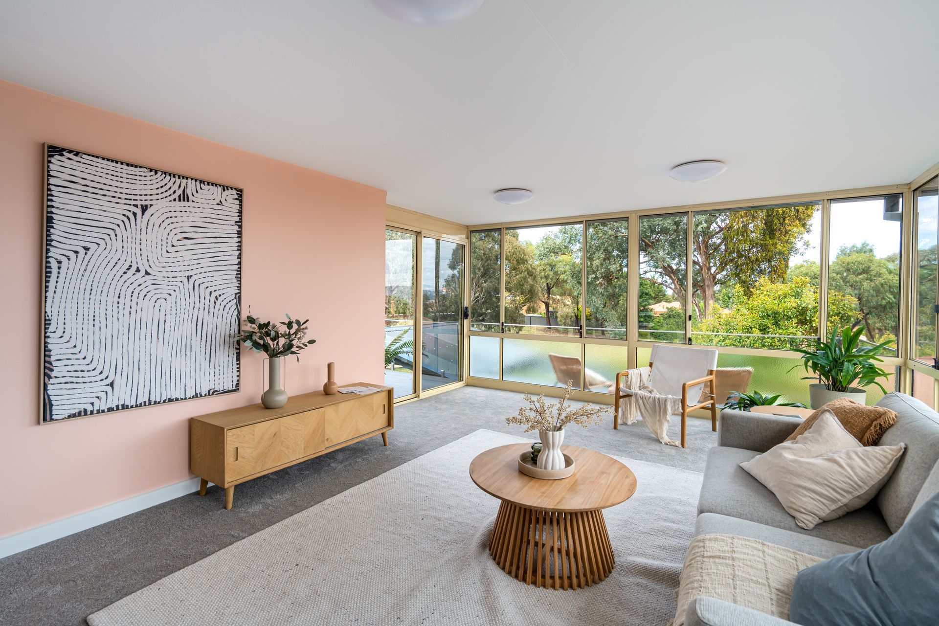 Living room with pink wall, wooden furniture, large windows overlooking a green yard, and a grey rug.