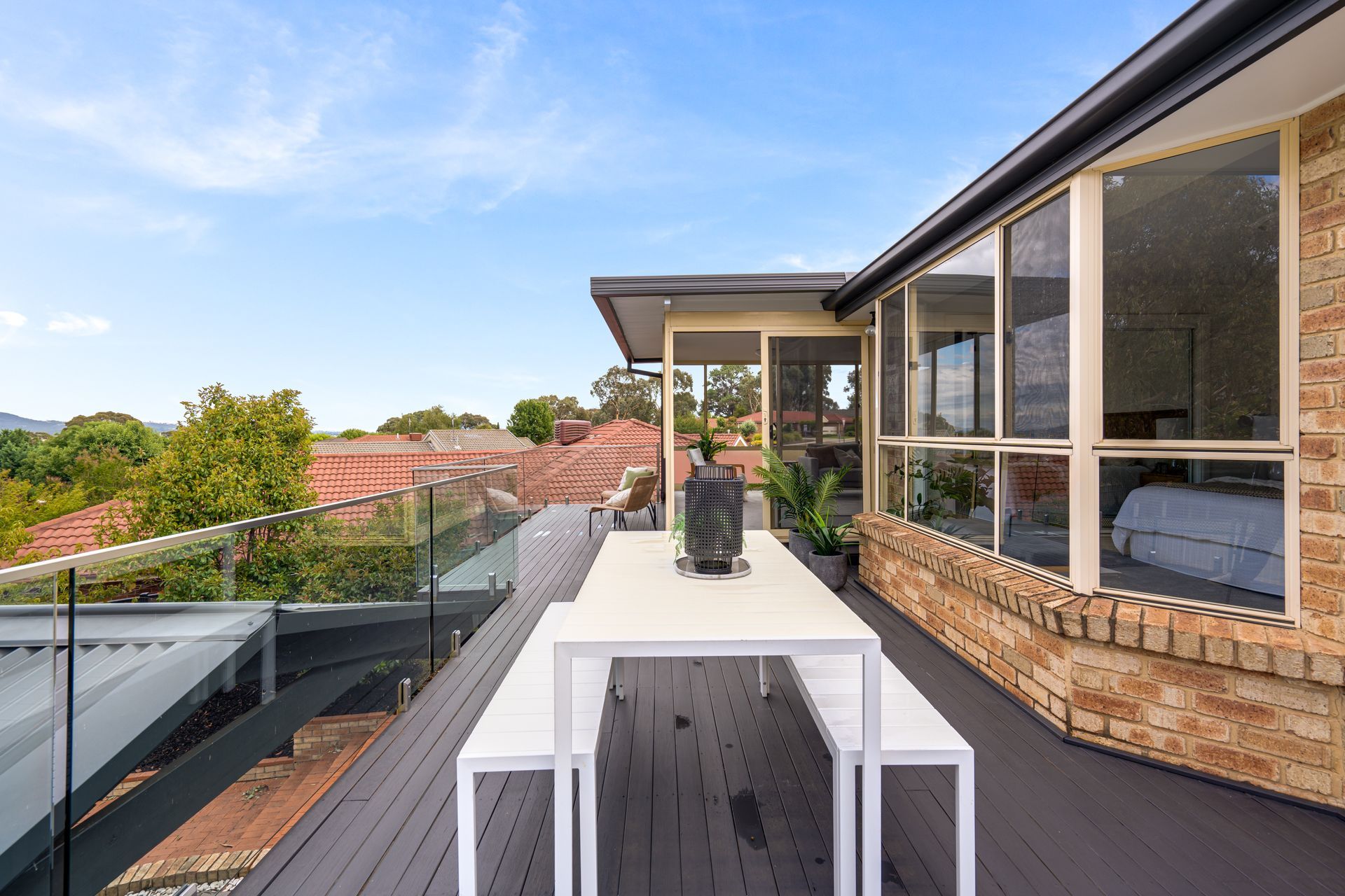 Outdoor deck with white table and benches, glass railing, brick wall, and view of rooftops under a blue sky.
