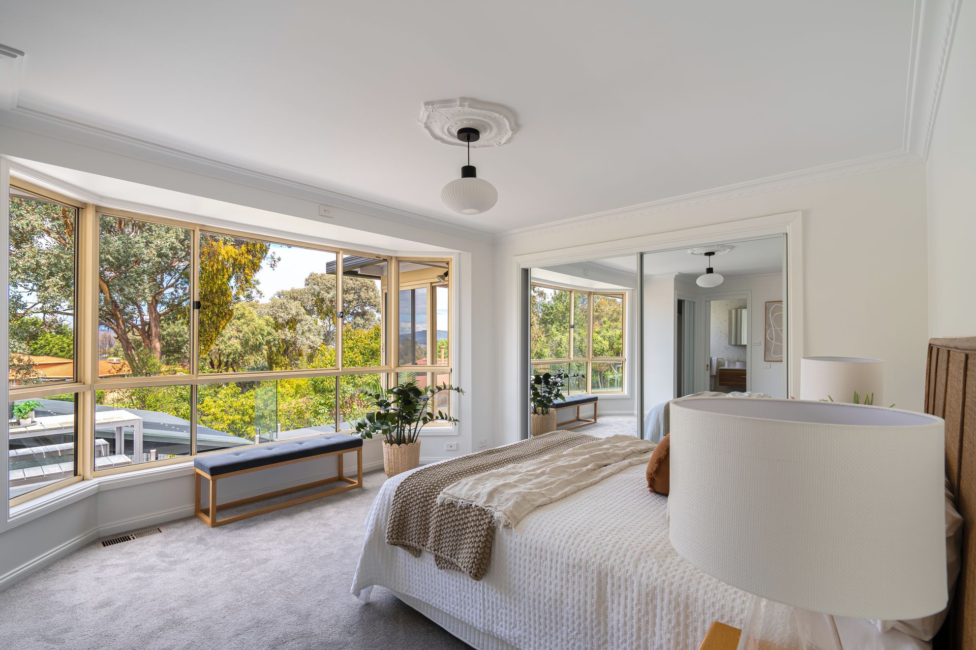 Bedroom with large window overlooking trees, a bed, bench, and a modern light fixture.