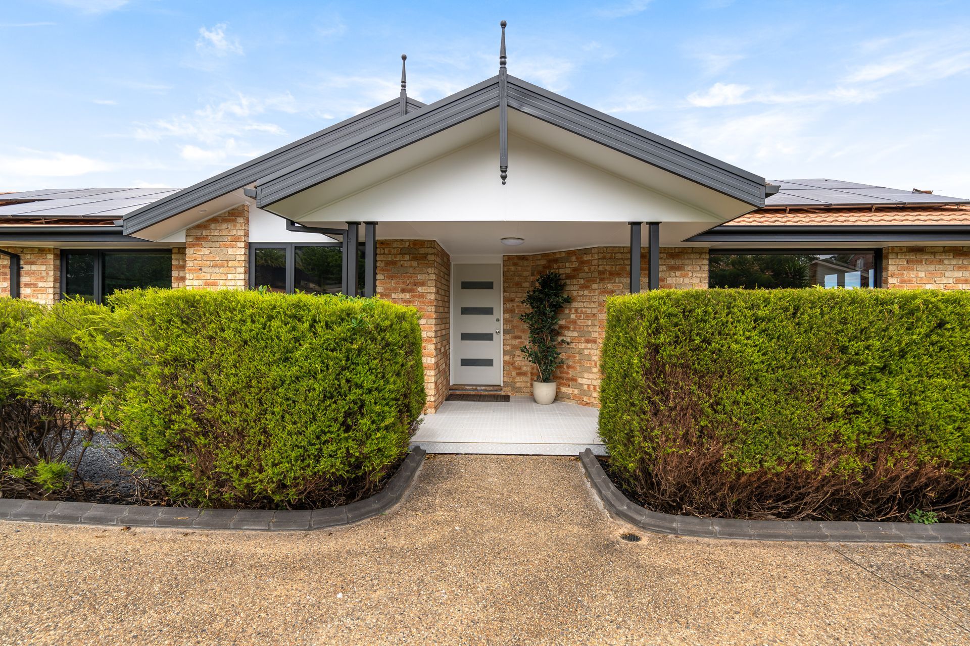 Exterior of a brick home with covered entrance, flanked by trimmed green hedges. Gravel path leads to the front door.