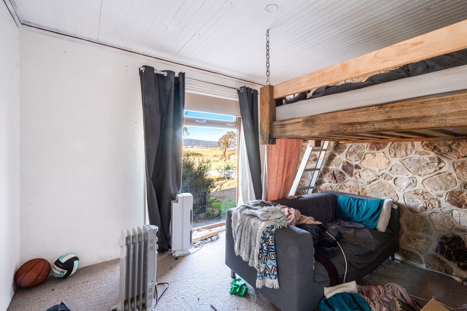 Interior of a rustic room with stone wall, couch, small loft, window overlooking a rural landscape.