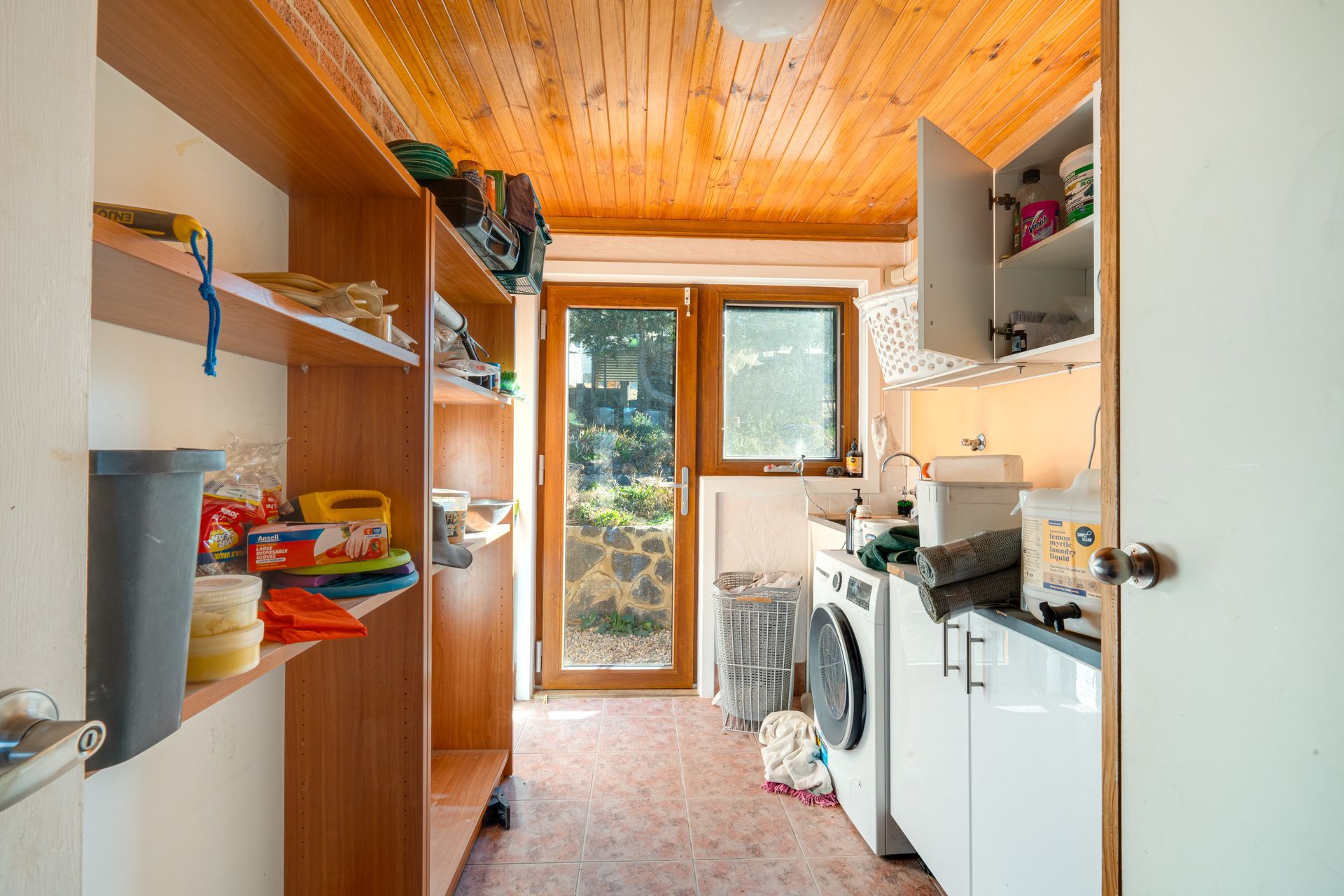 Laundry room with washer, dryer, shelves, and window looking out to greenery. Wooden ceiling and cabinet.