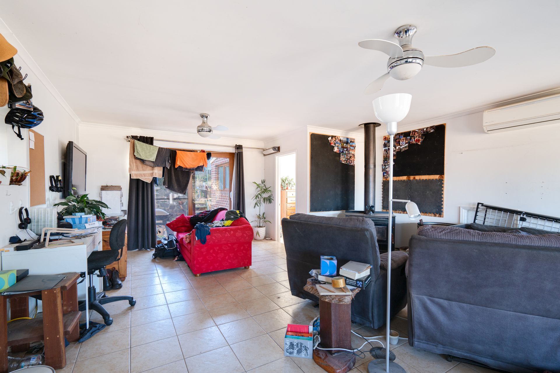 Spacious living room with assorted furniture, cluttered with clothes and objects. Ceiling fan and floor lamp provide lighting.