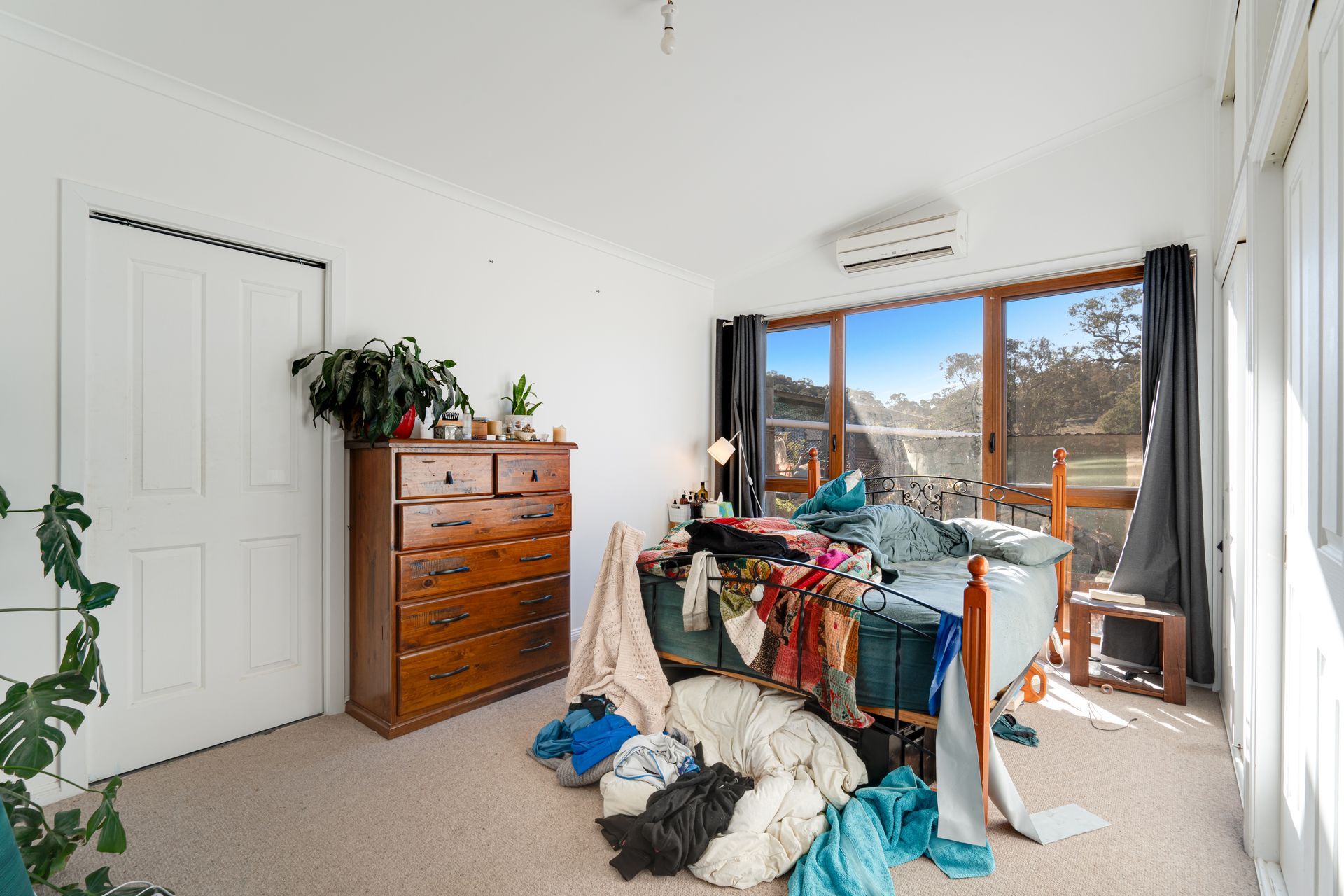 Messy bedroom with clothes strewn about, unmade bed, wooden dresser, window with bright view.