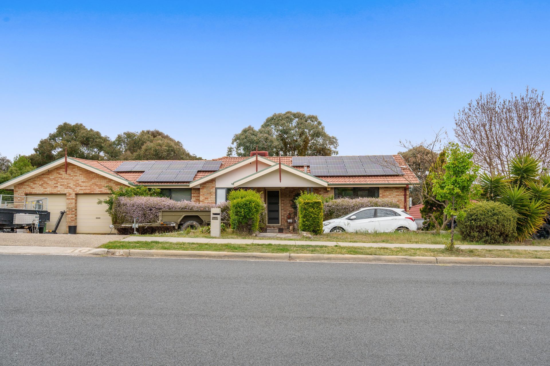 A single-story brick house with solar panels, a garage, a car, and a trailer on a suburban street.