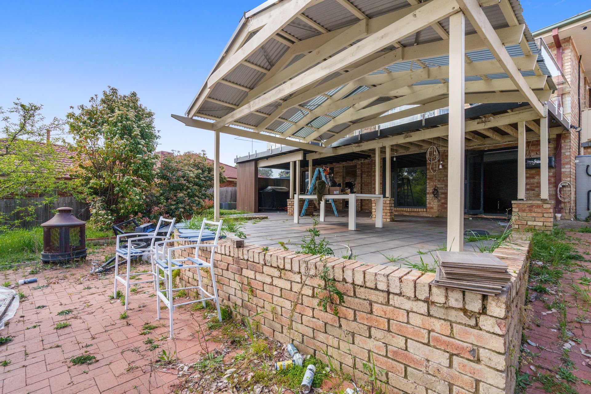 Backyard patio with brick wall, overhead shelter, chairs, and overgrown weeds.