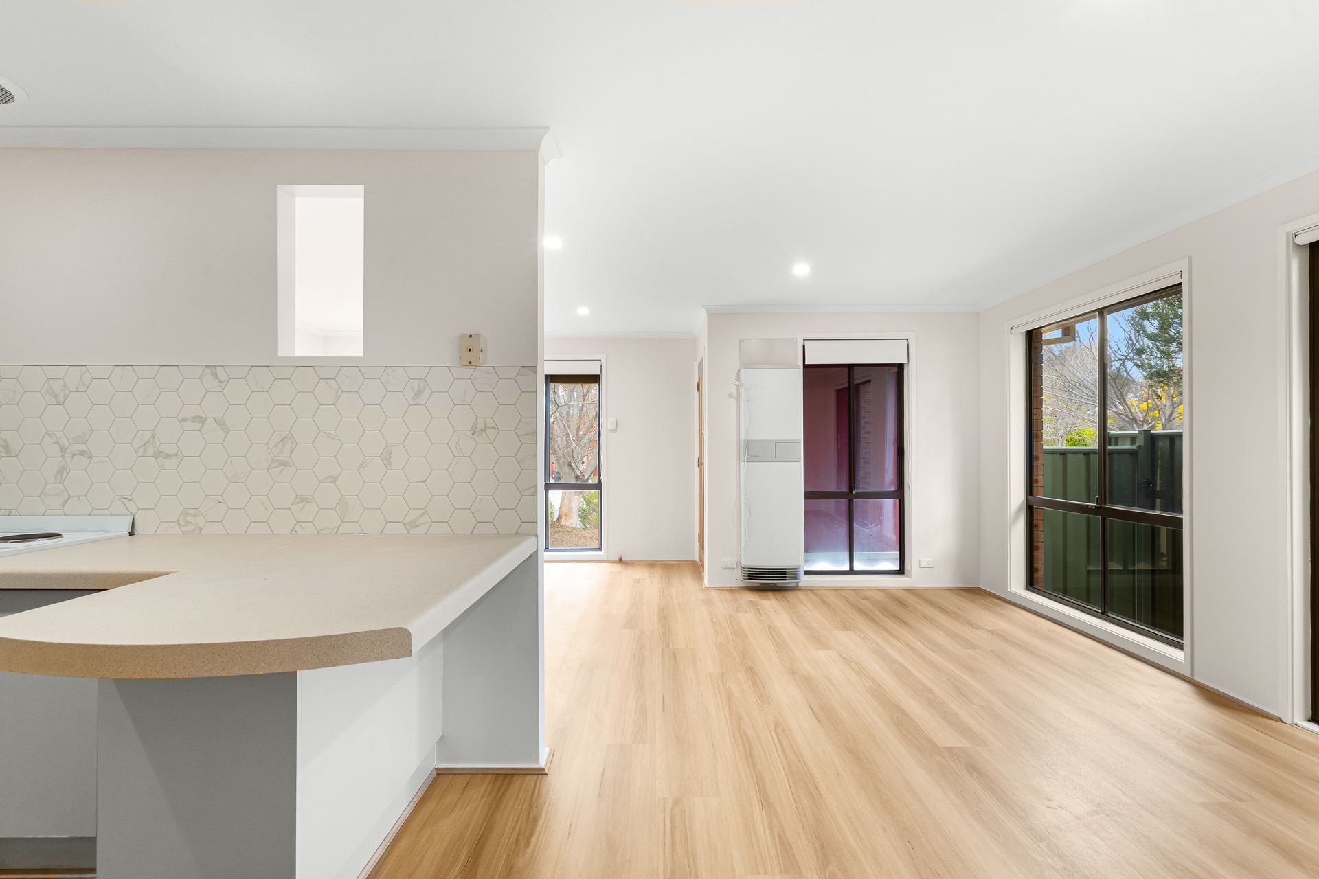 Interior of a bright, empty living space with wood-look flooring, windows, and kitchen counter.