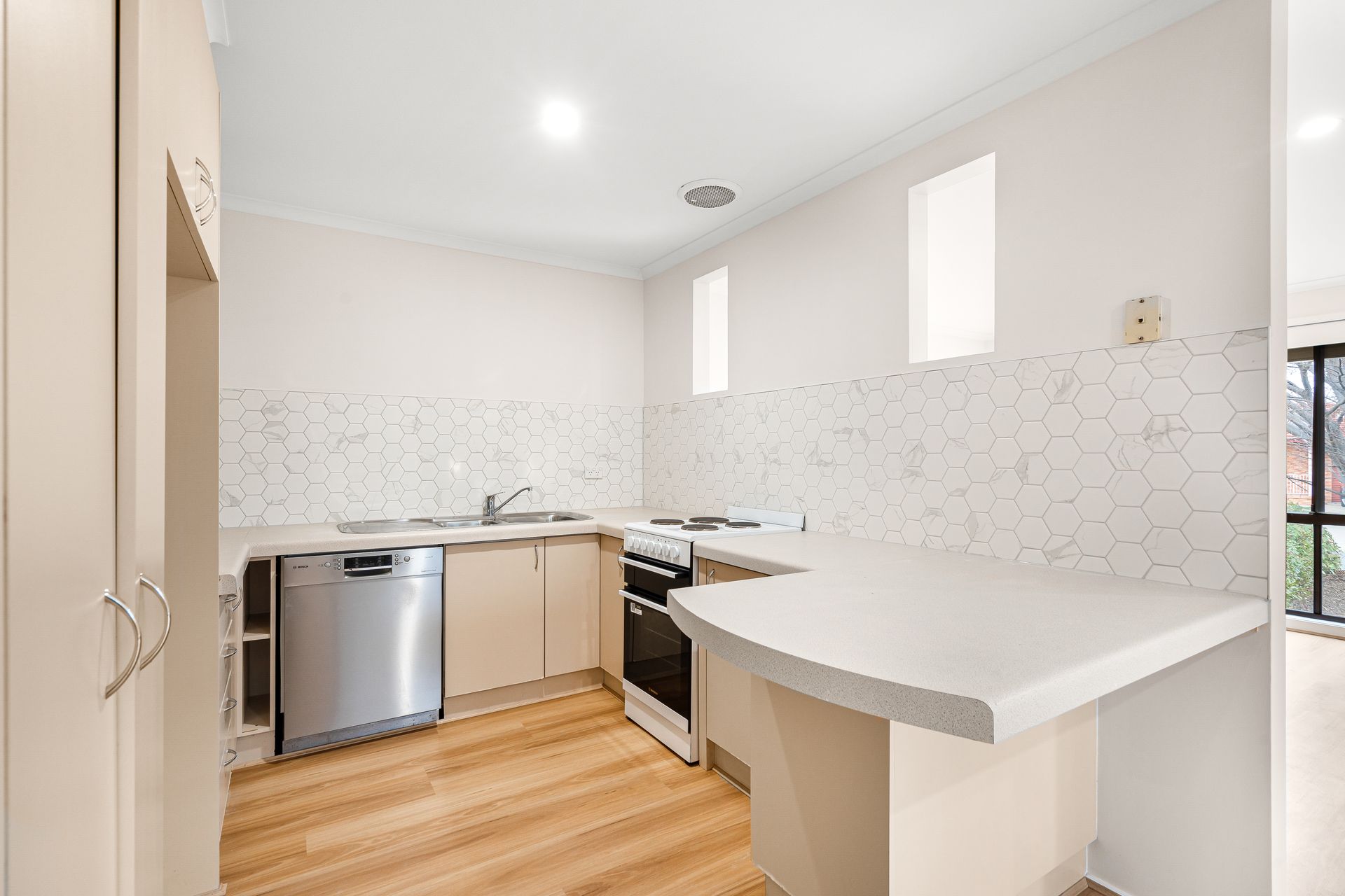 Kitchen with light wood floor, beige cabinets, white countertop, backsplash, and appliances.