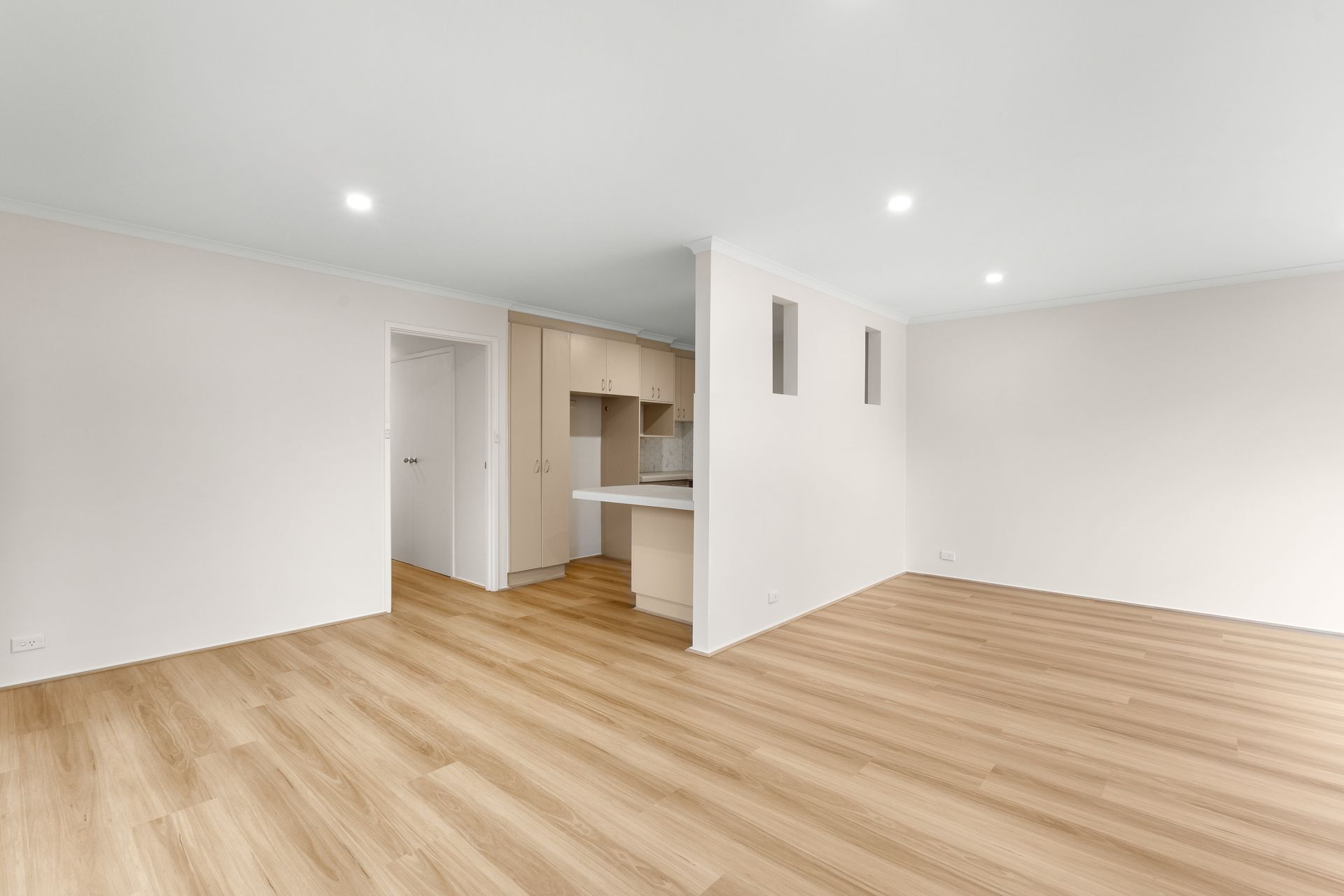 Empty modern apartment interior, light wood floors, white walls, kitchen in background.