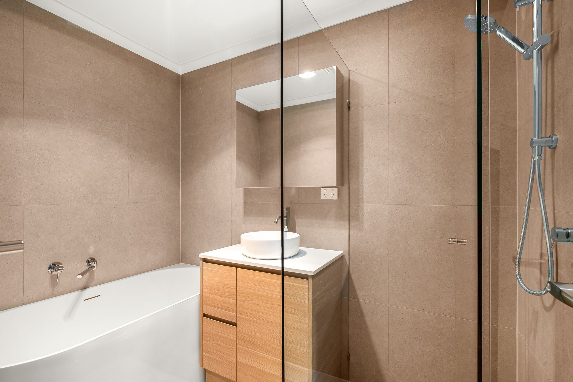 Bathroom with tan tile walls, white bathtub, wooden vanity, and glass shower door.