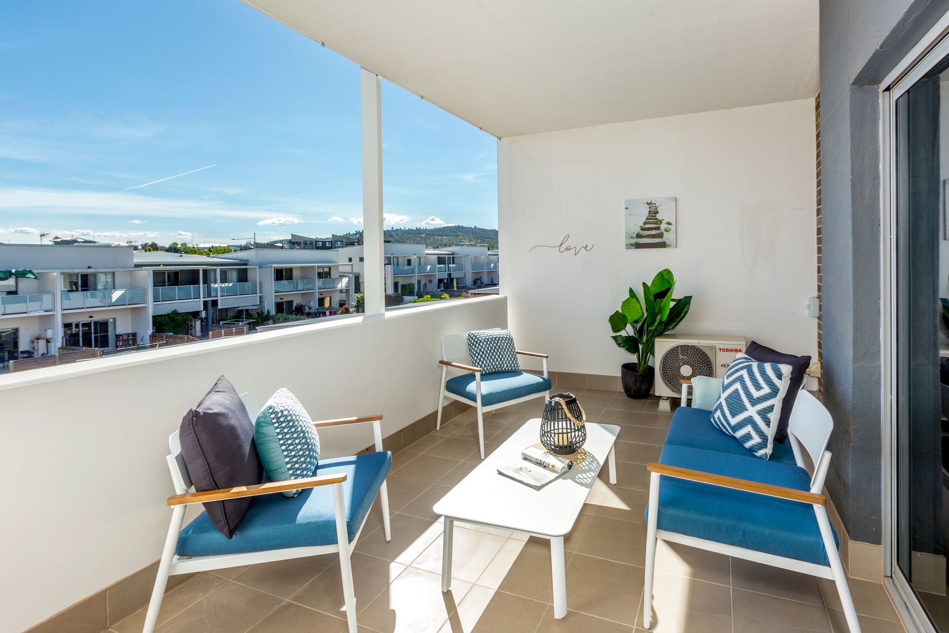 Balcony with blue seating, white table, and potted plant, overlooking a residential area on a sunny day.