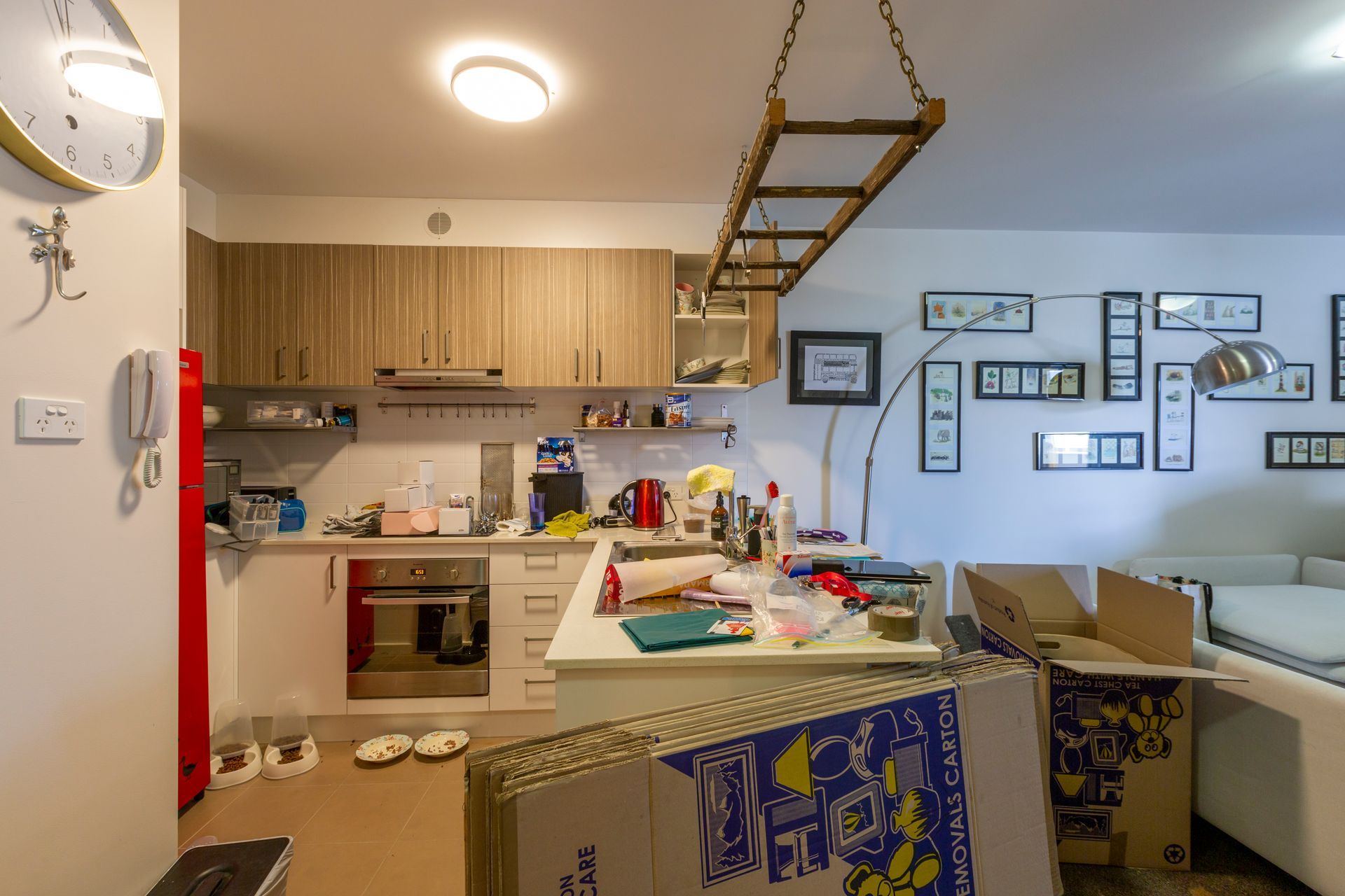 Kitchen with island and cabinets. Boxes are in front of the island. Wall art and couch visible.