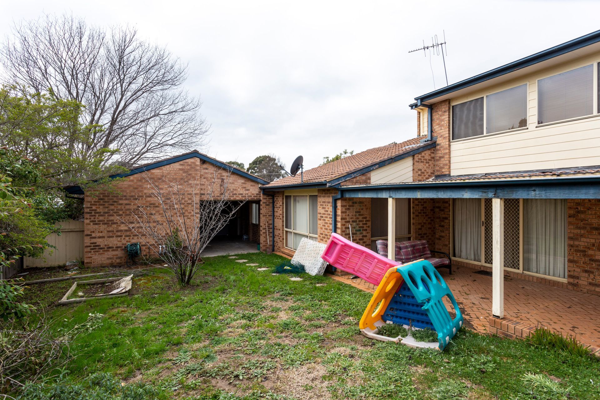 Backyard with brick house, detached garage, and children's play equipment on a grassy lawn under a cloudy sky.