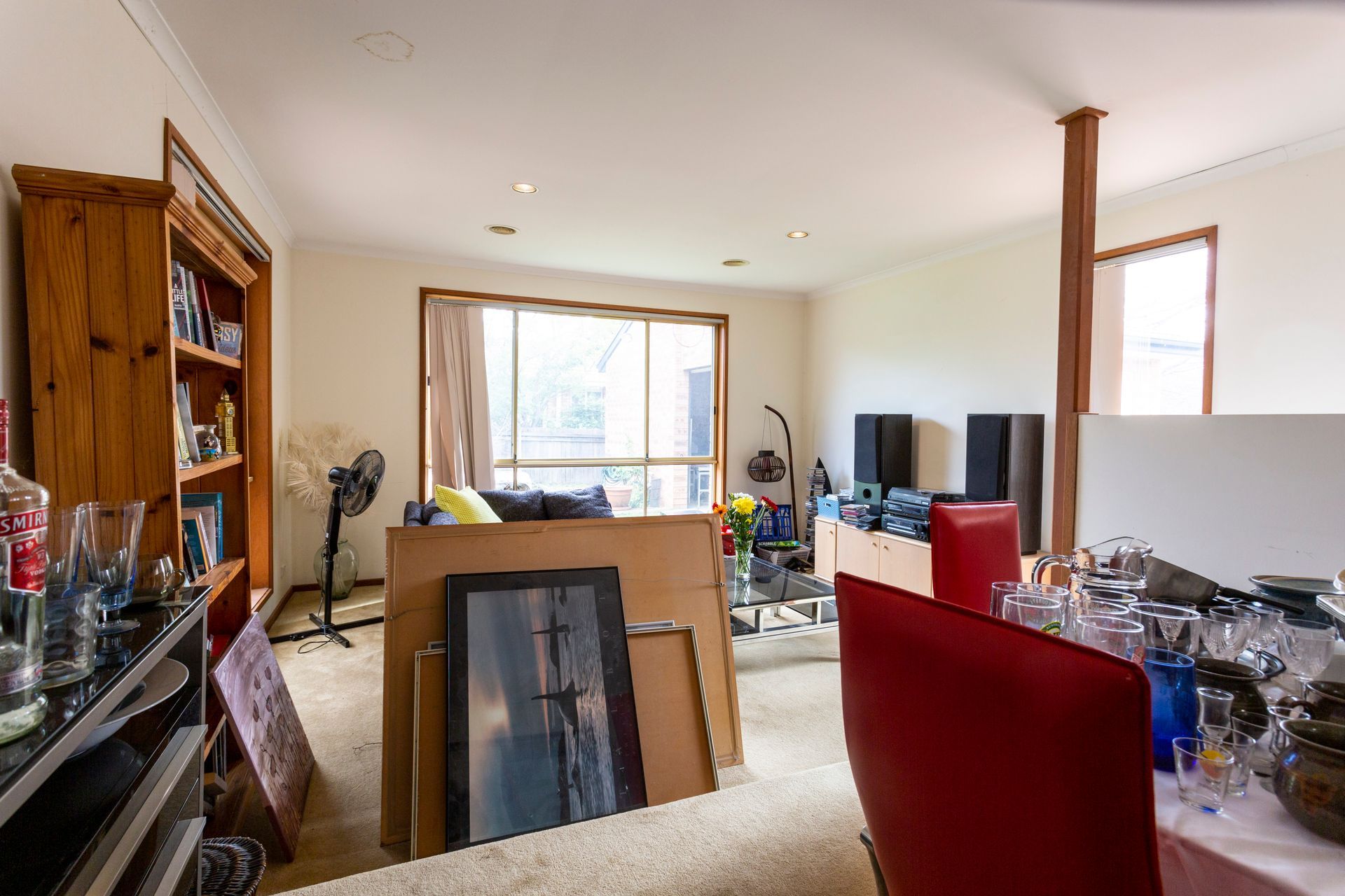 Cluttered living room with furniture, boxes, and a wooden bookshelf near a window.