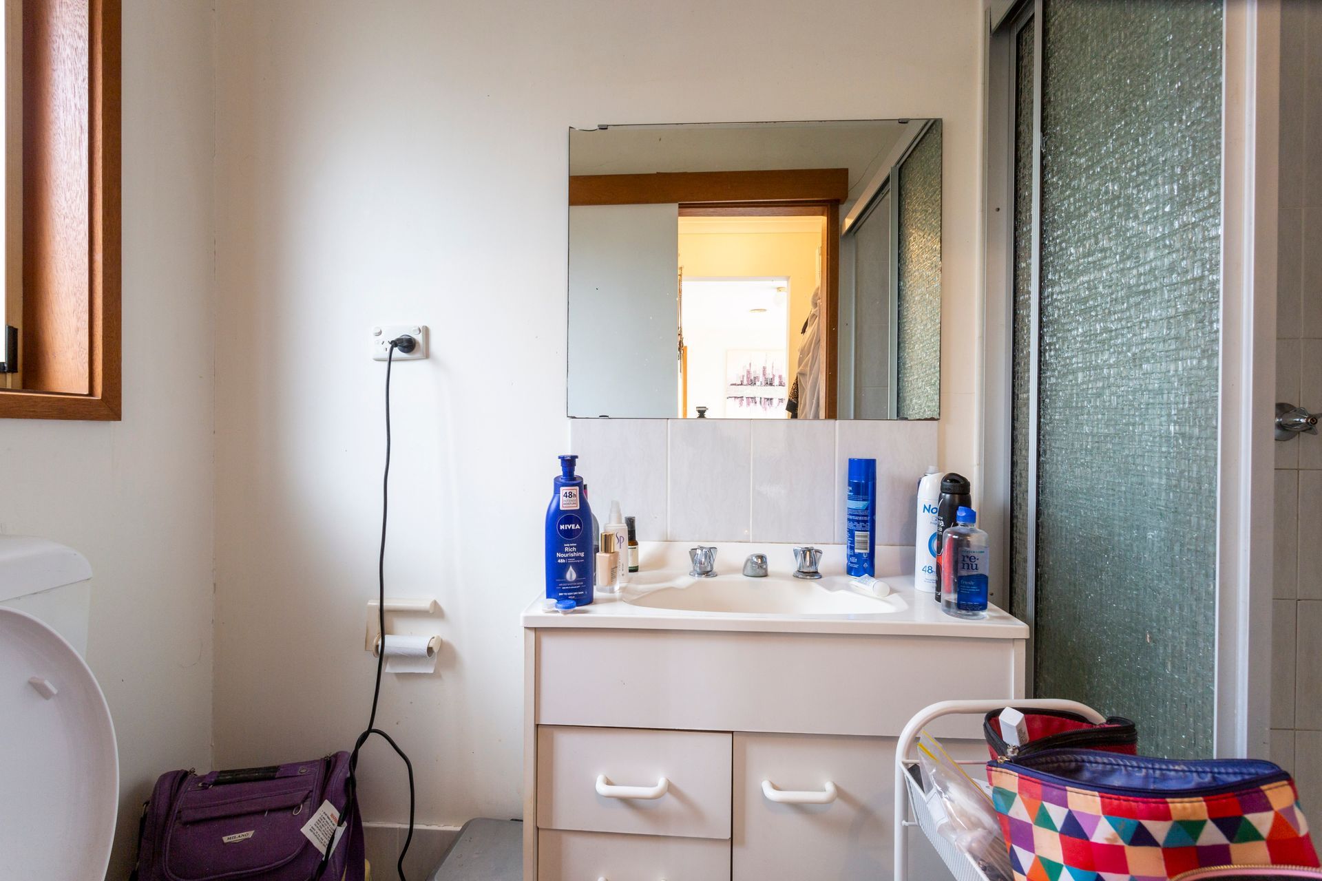 Bathroom with sink, mirror, shower, and luggage; various toiletries on the counter.