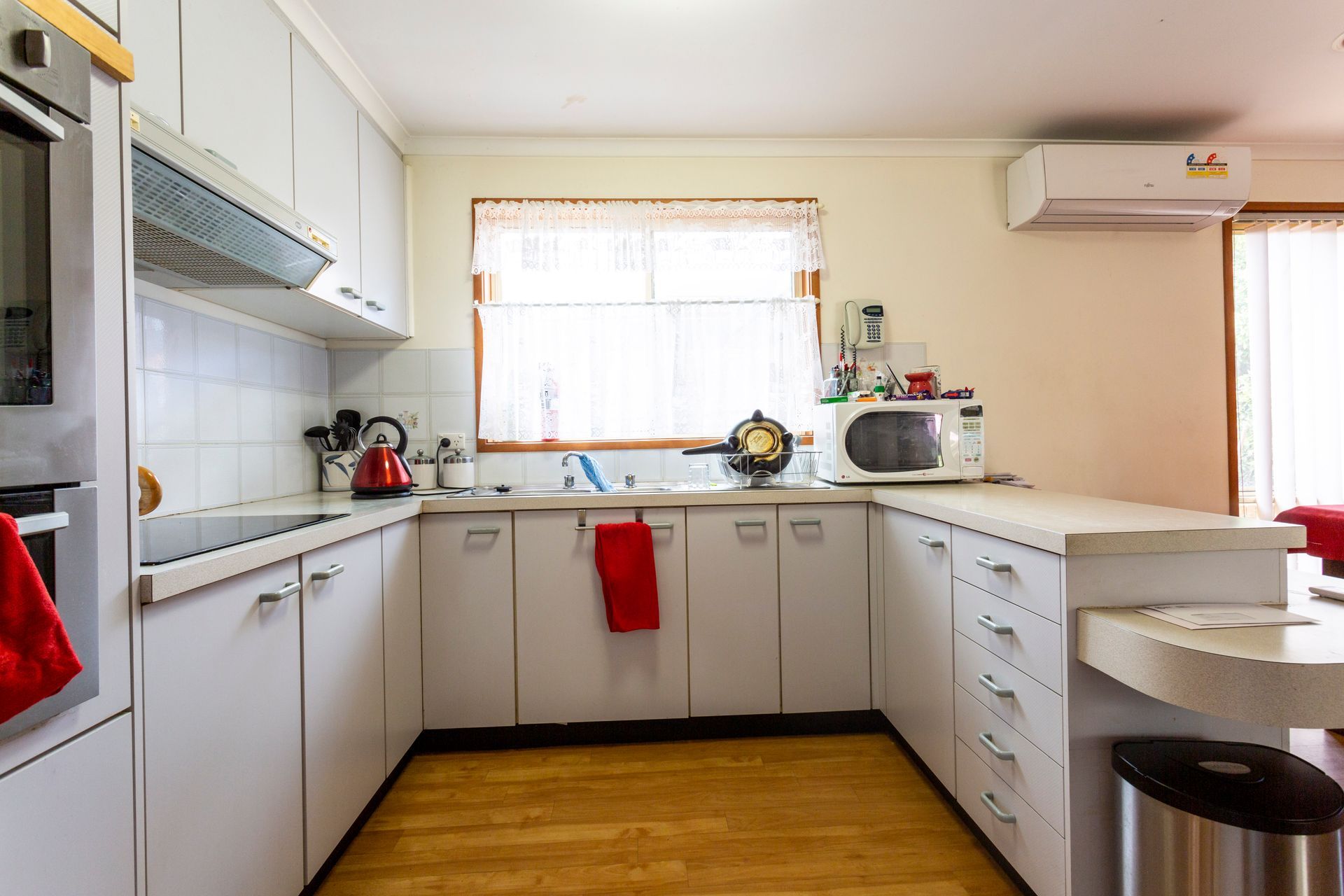 White kitchen with cabinets, stove, microwave, and window with sheer curtains. Wooden floors.