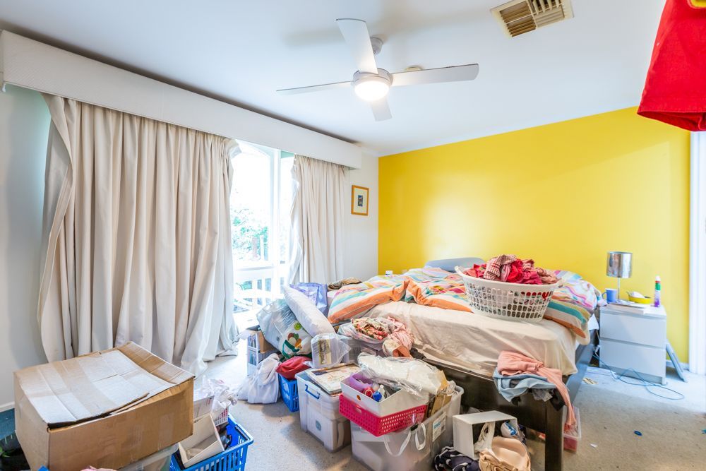 Messy bedroom with wardrobe, dresser covered in clothes, fan, and cluttered floor.