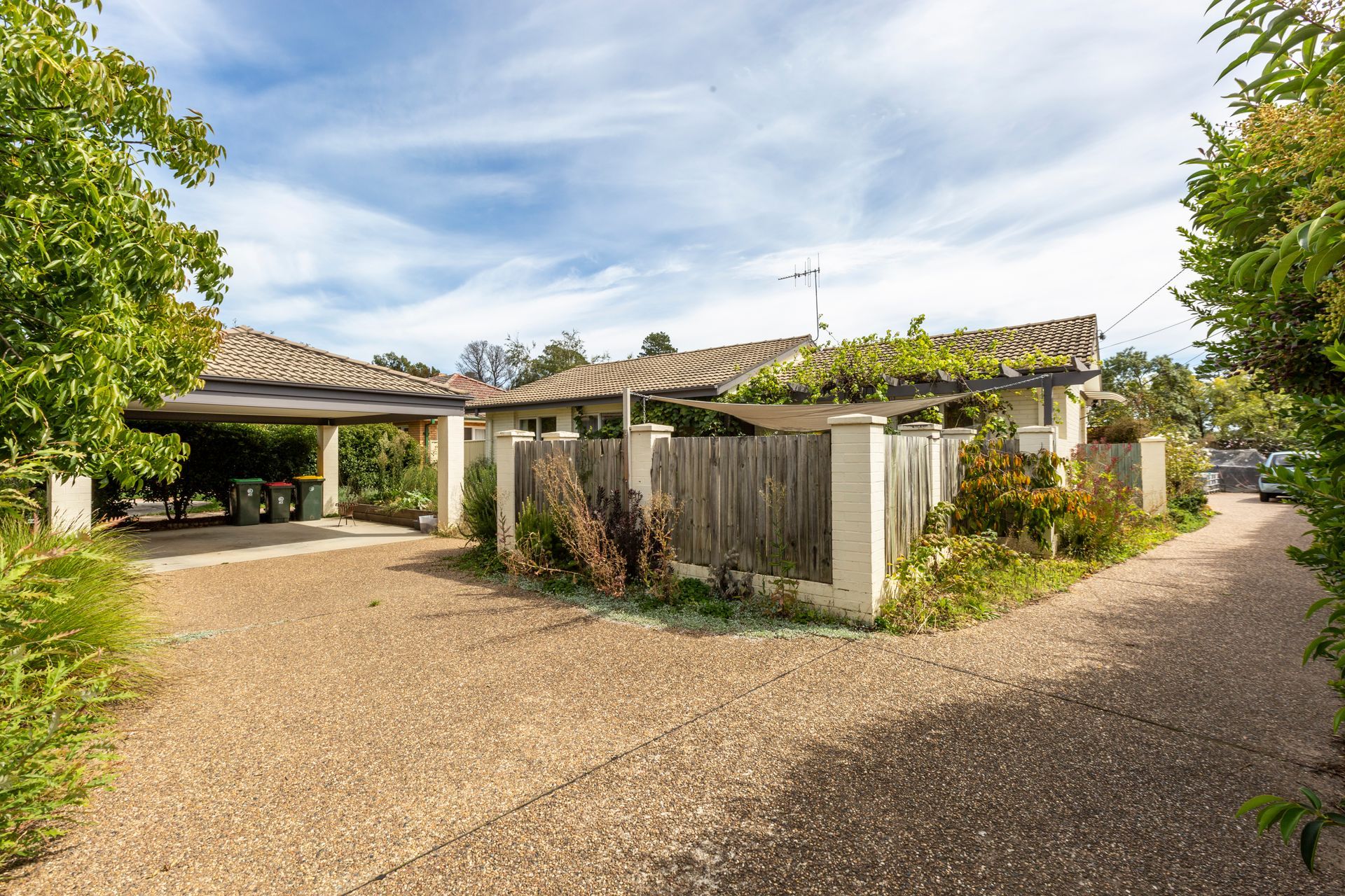 Beige house with gravel driveway, carport, and wooden fence under a cloudy sky.